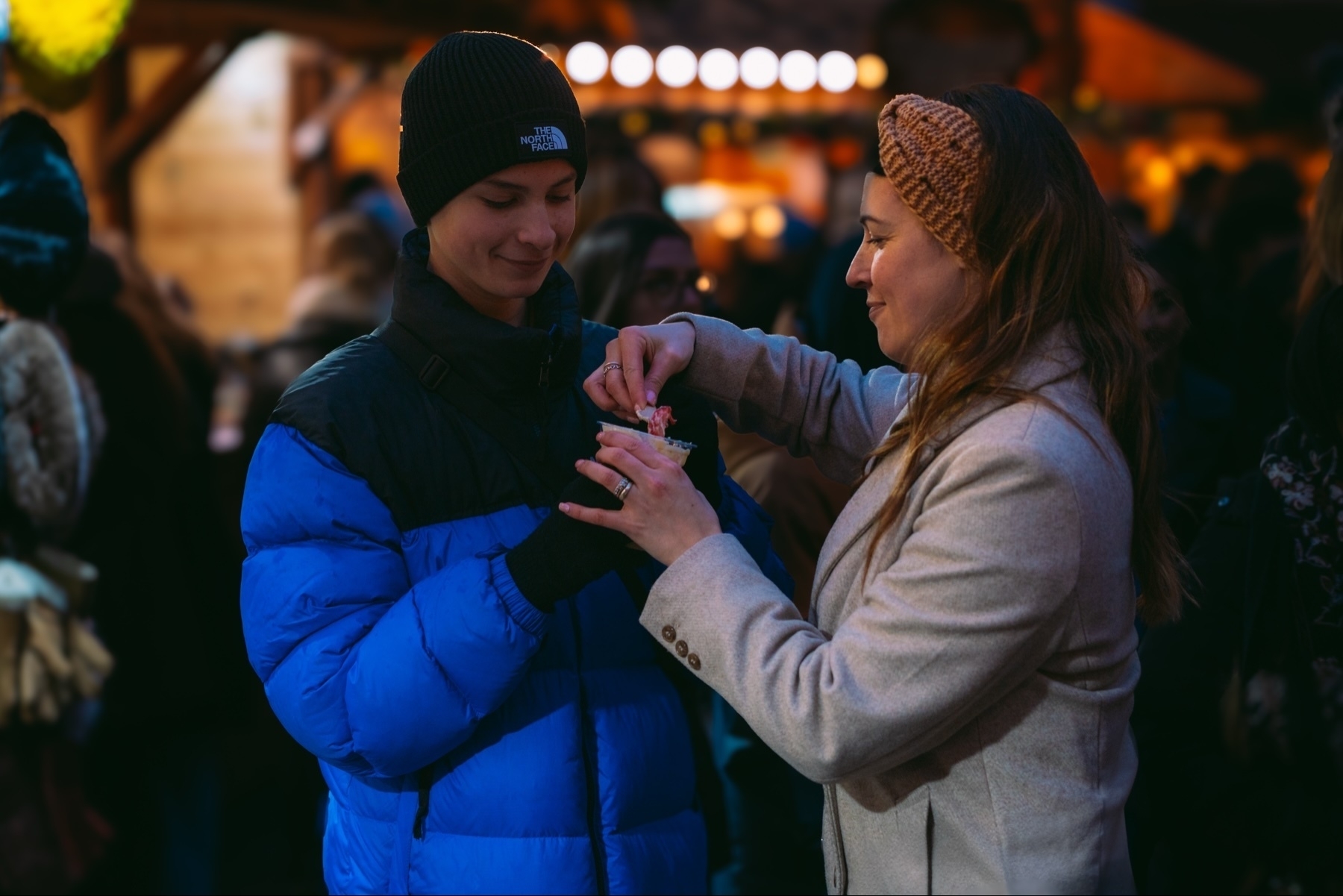 A person in a blue jacket and black beanie is holding a cup while another person in a light coat and brown headband is reaching into it. They are in a crowded, warmly lit marketplace setting.