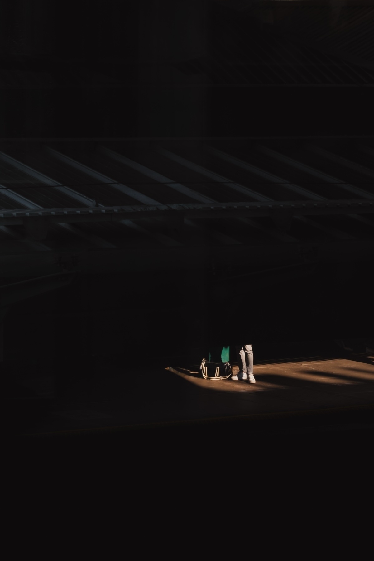 A person stands in a spotlight under a dark, possibly industrial or architectural structure. Only their lower half is visible, and they have a suitcase or bag beside them. The lighting creates a dramatic contrast with the surrounding darkness.