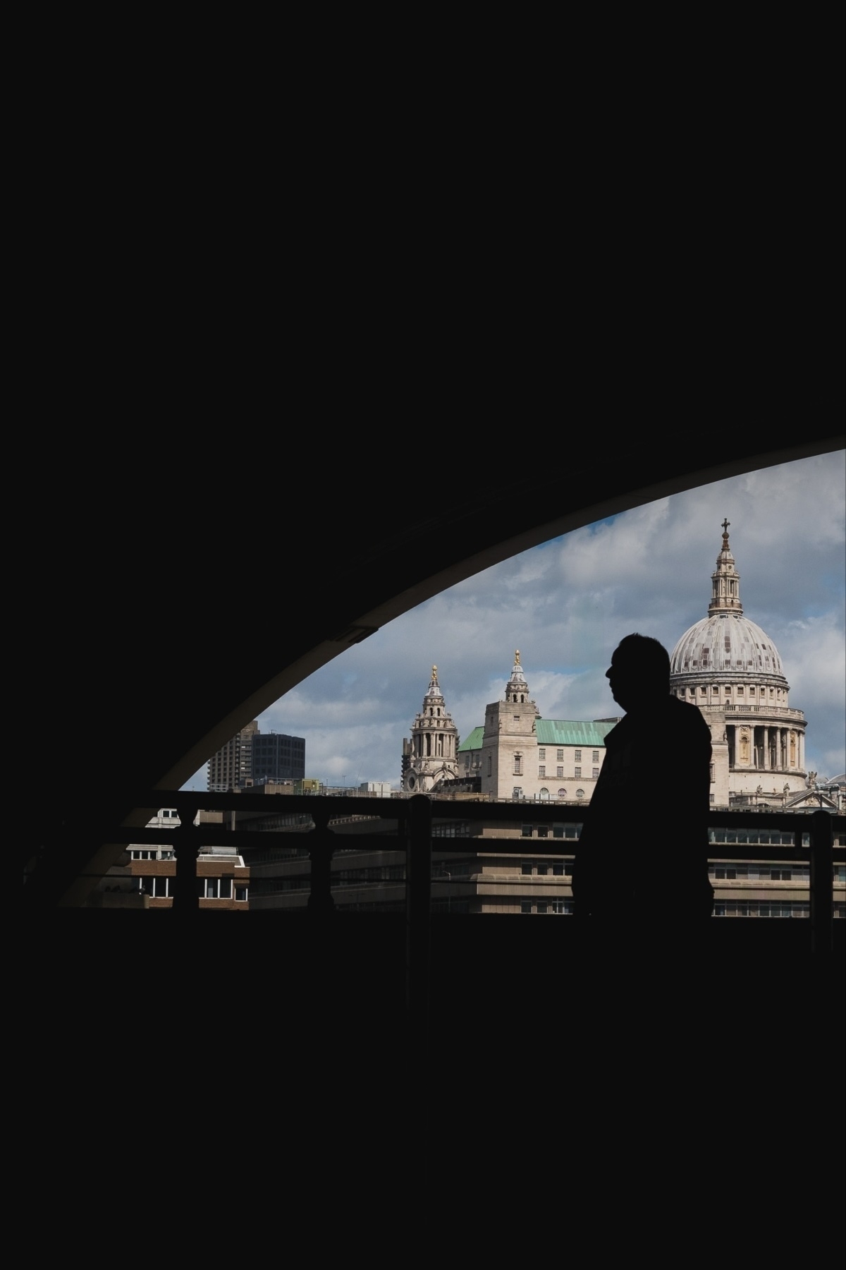 A silhouette of a person standing in front of a railing, with a view of St. Paul's Cathedral and other buildings in the background. The image is framed by a dark arch.