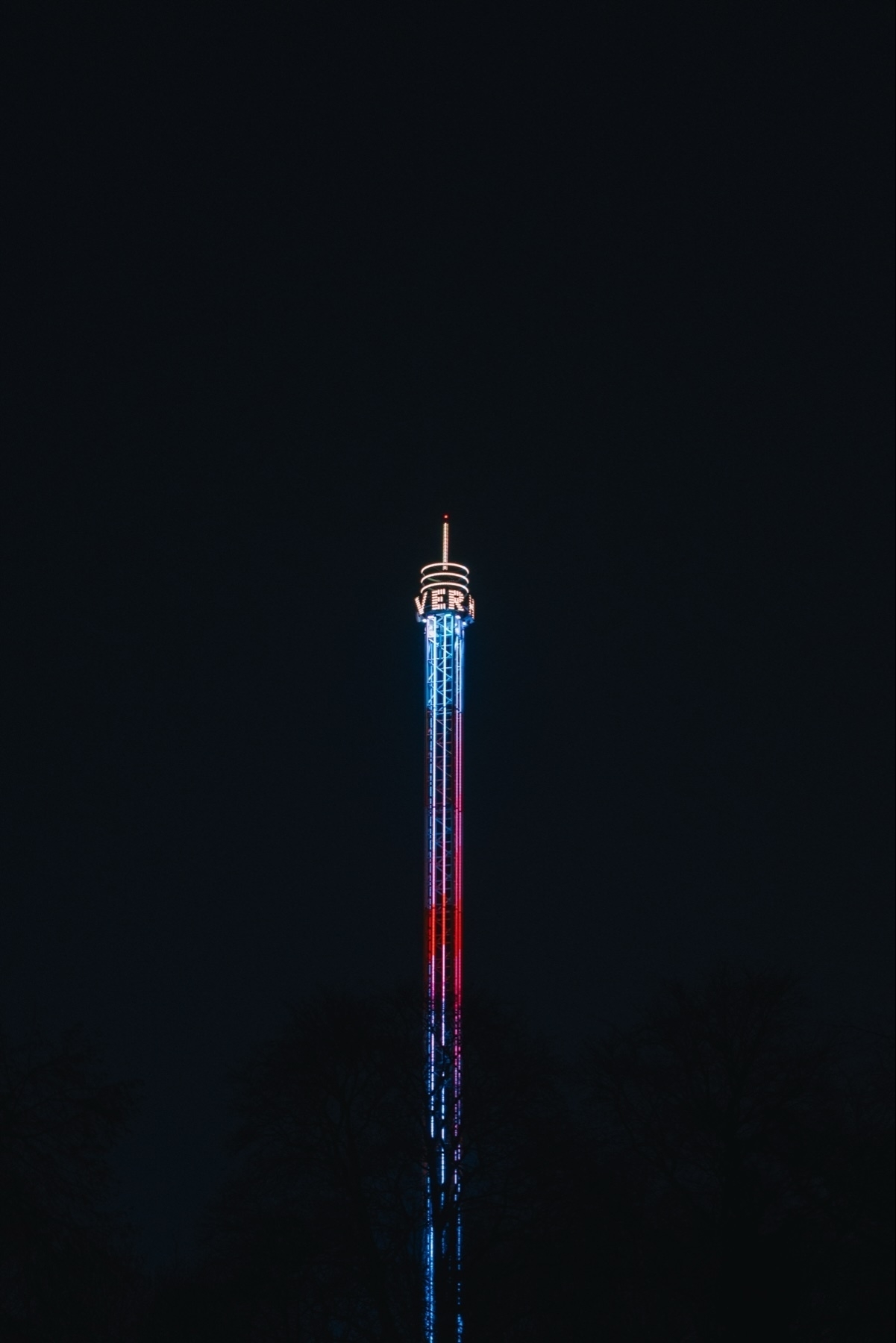 A tall illuminated tower at night with vertical red and blue lights, surrounded by silhouettes of trees at the base. The top of the tower displays the word 