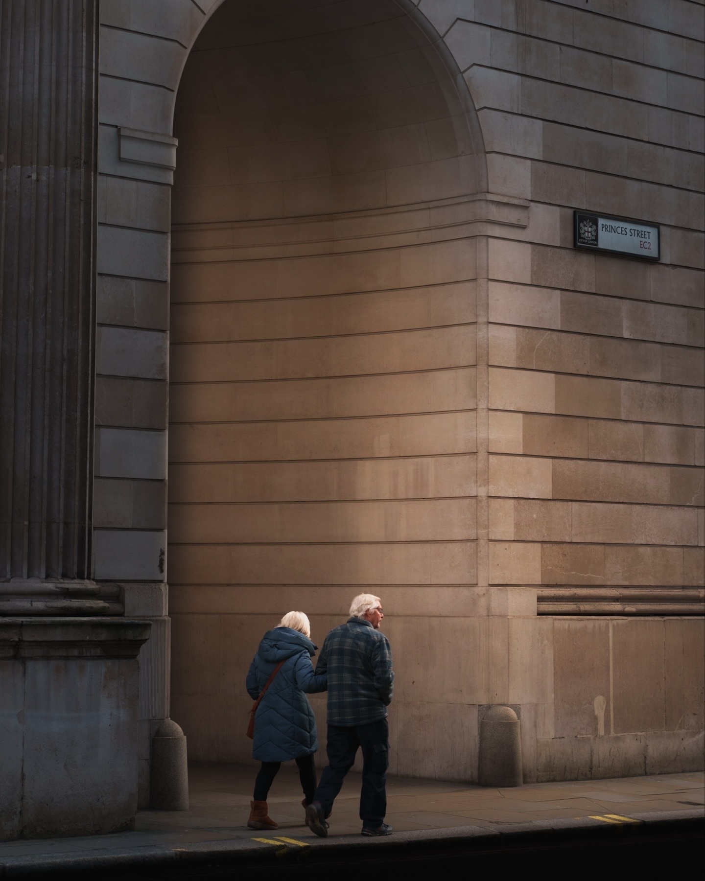 An elderly couple walks hand in hand along a sidewalk near a large stone building with an archway. The building has a sign reading “Princes Street EC2.” The scene is calm and the lighting is soft.