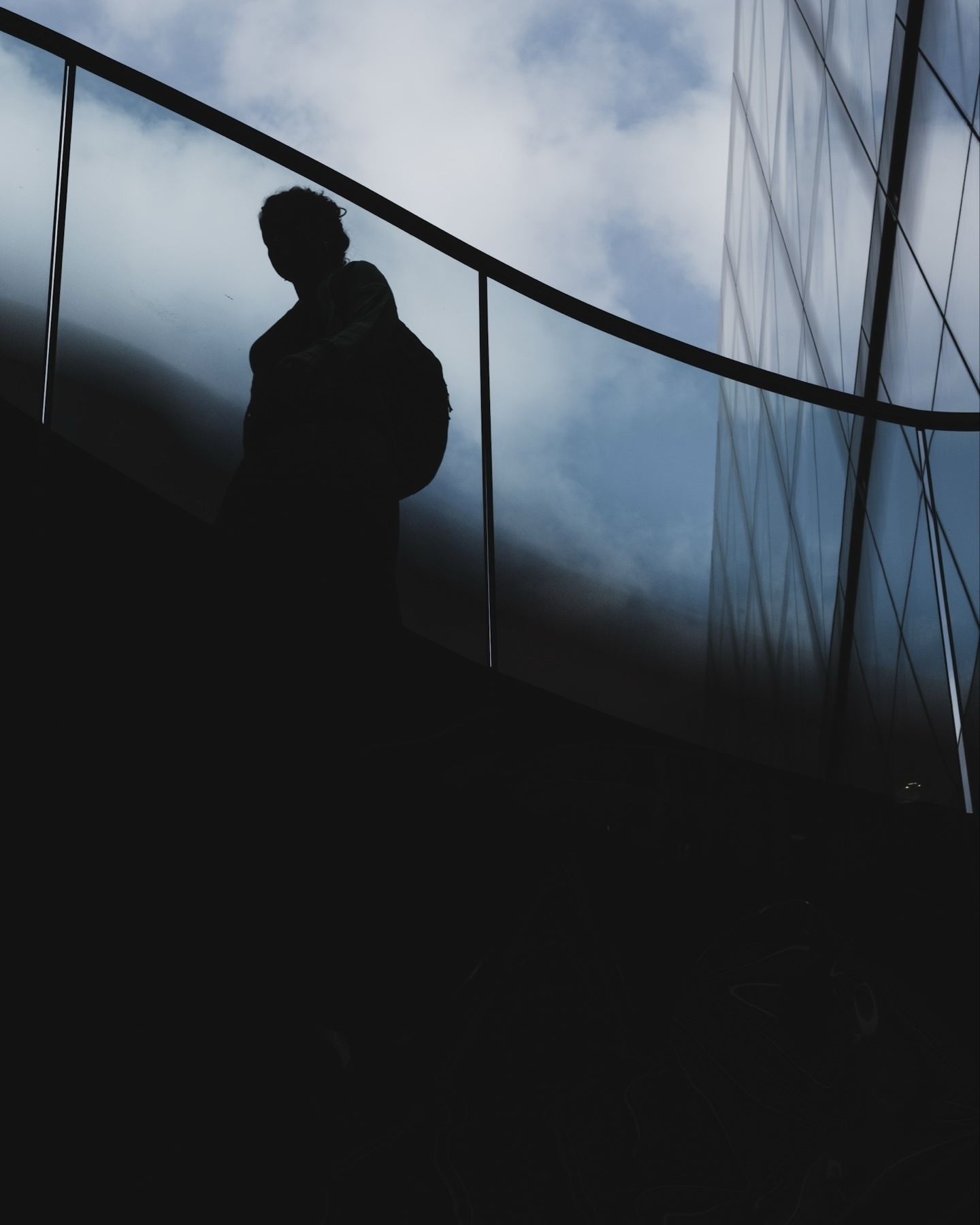 Silhouette of a person with a backpack on a curving staircase, set against a backdrop of glass and metal architecture under a cloudy sky.