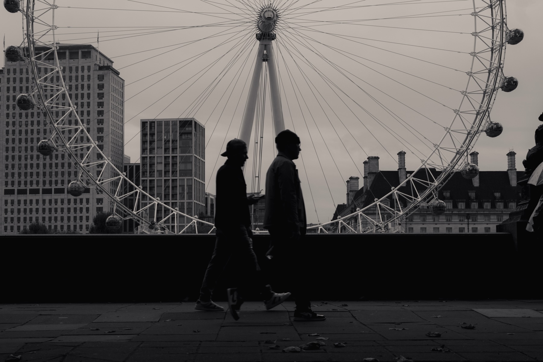 Silhouette of two people walking in front of the London Eye ferris wheel. The scene is set in an urban environment with buildings in the background, in a monochrome color scheme.