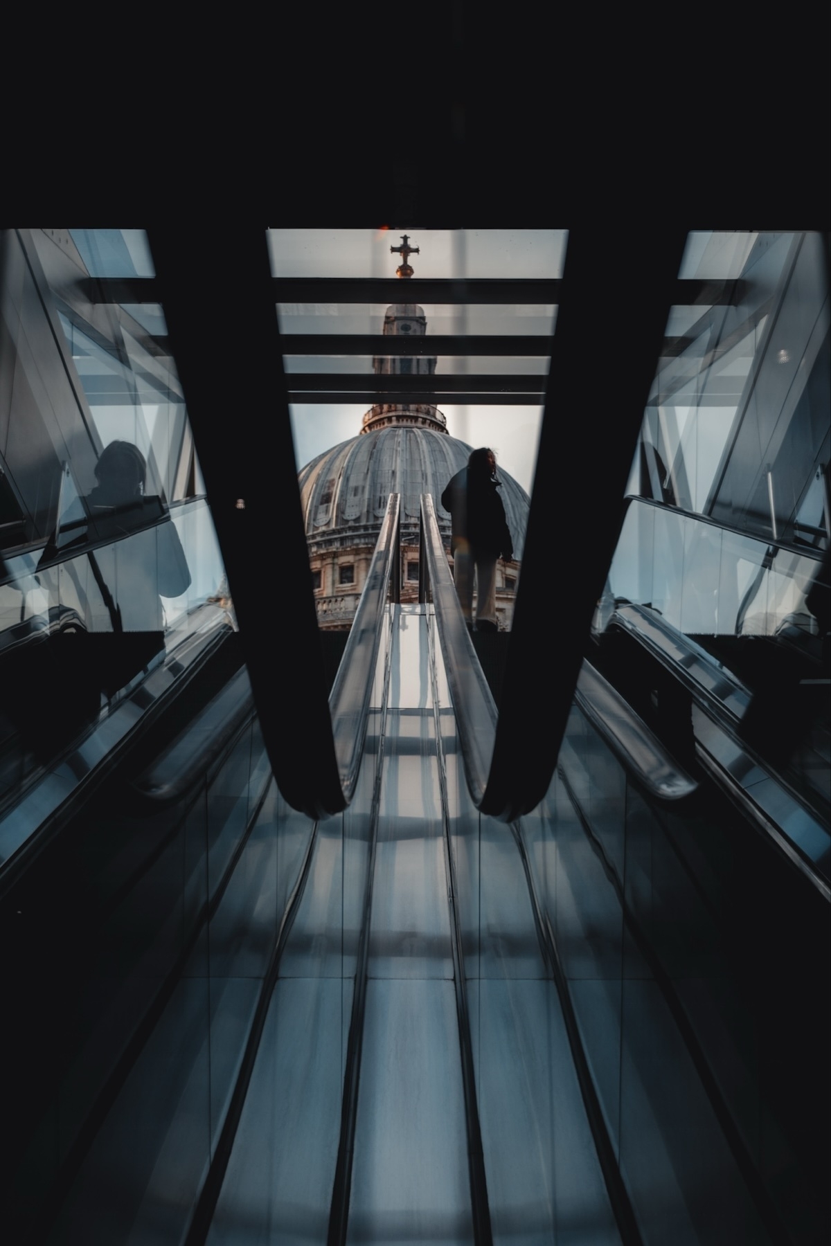 View of an escalator inside a building with a silhouette of a person ascending. In the background, the dome of St. Paul’s Cathedral is visible through glass panels. The scene is framed by the dark interior, creating a dramatic contrast with the outside light