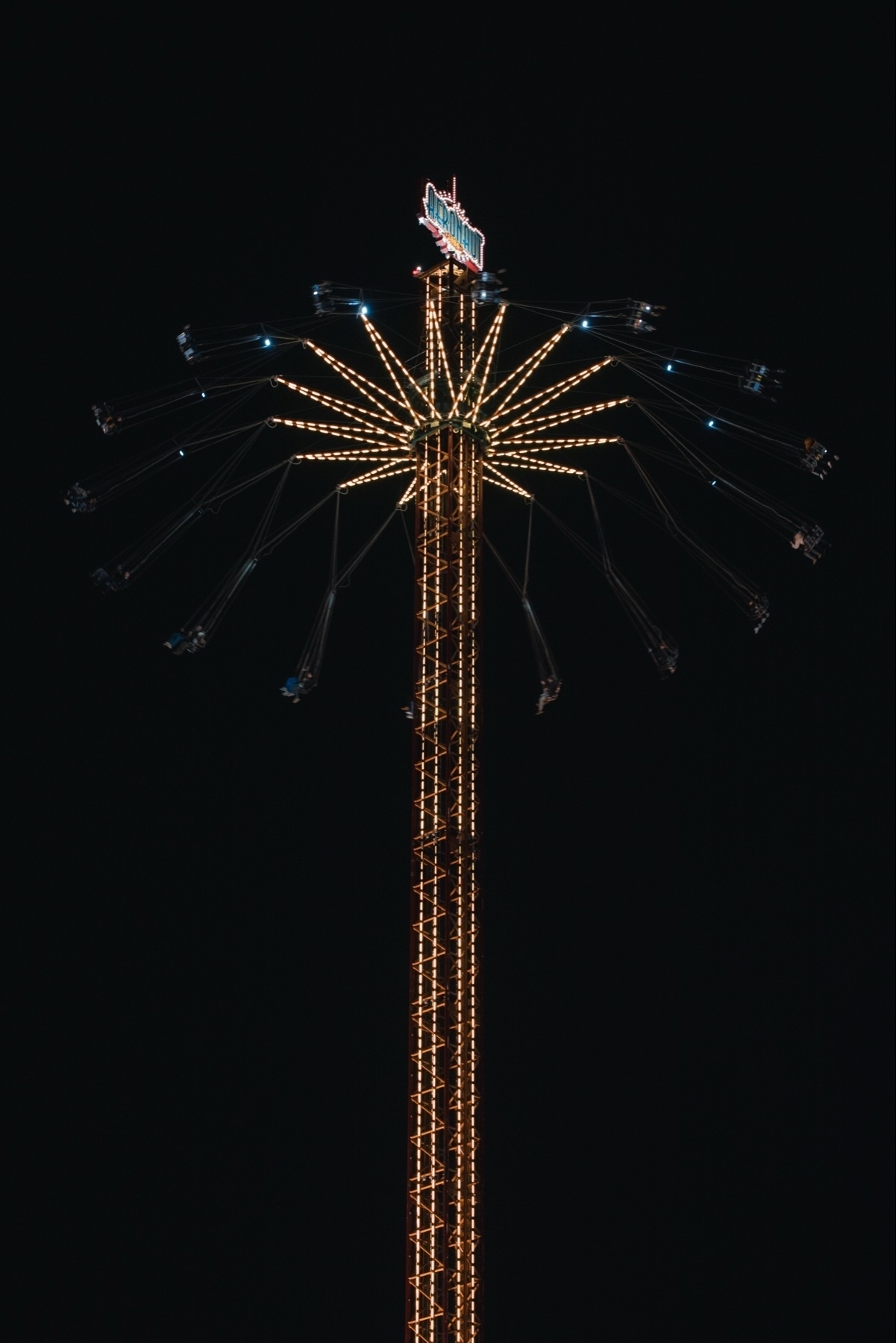 A tall amusement park ride lit up at night, resembling a star-shaped structure with swings extending outward. The dark sky serves as a backdrop.