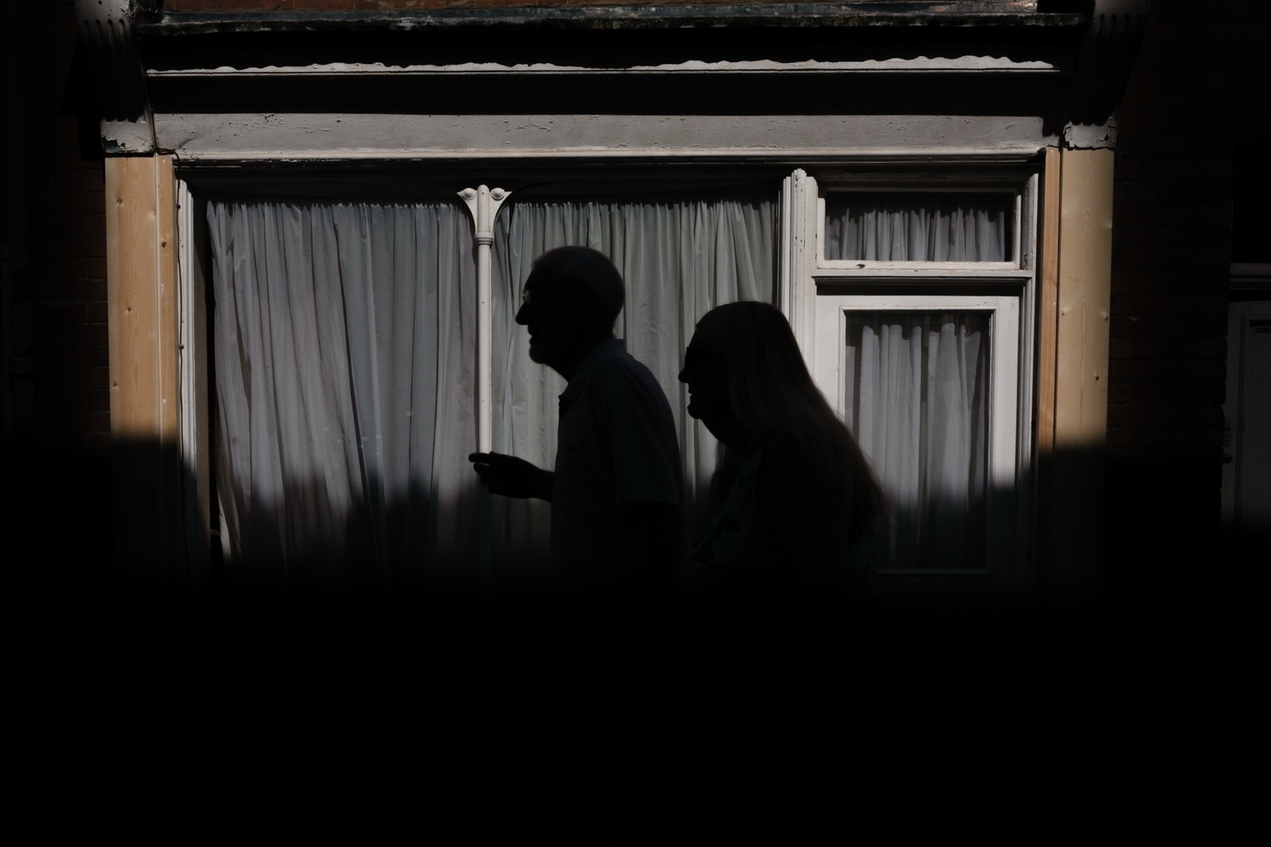 Silhouettes of a man and a woman walking past a window with closed curtains in the background. The image has strong contrast with light coming from above, creating a shadowed foreground.