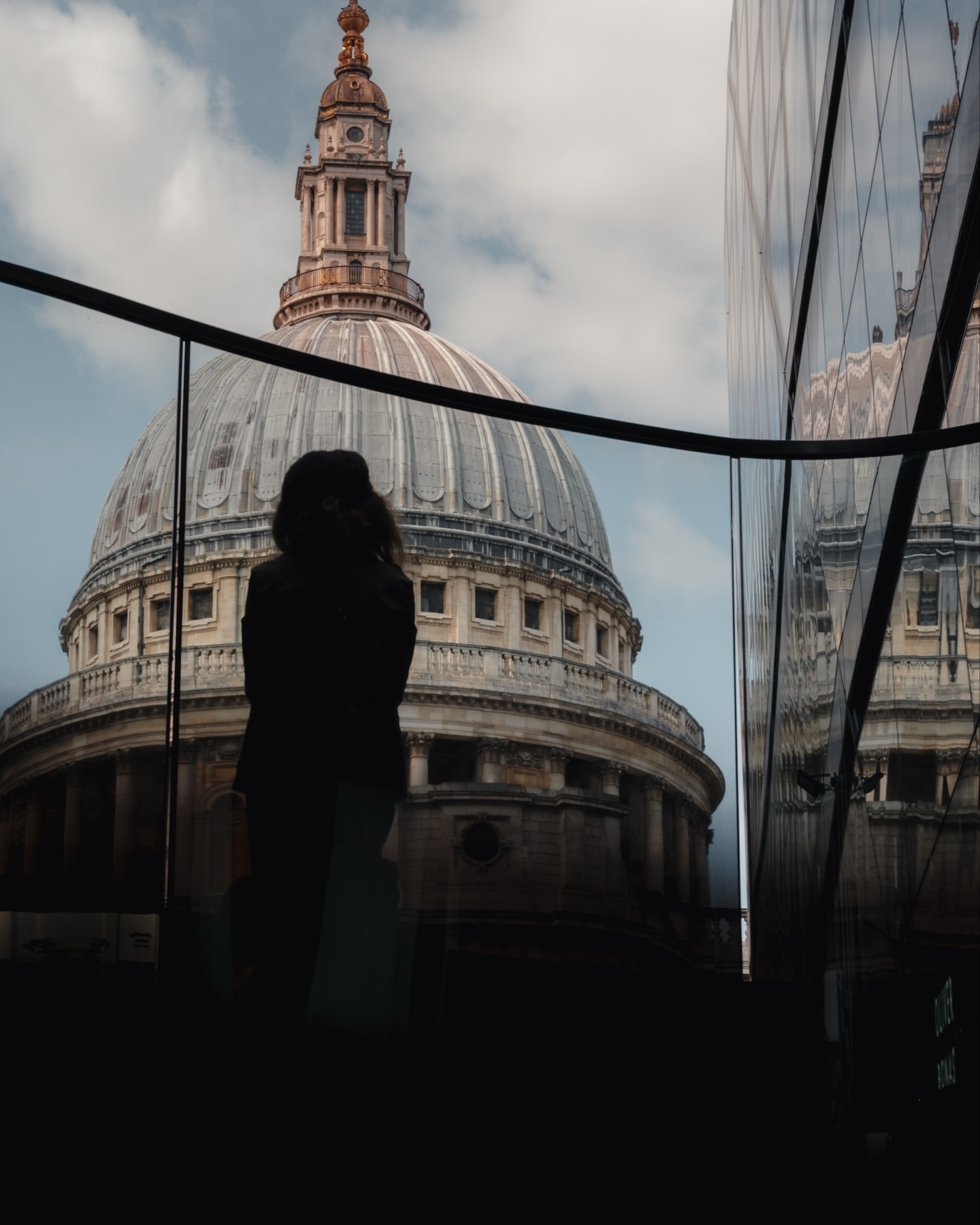 A silhouette of a person standing near reflective glass, with the dome of St. Paul's Cathedral in London prominently visible in the background. The glass also mirrors the cathedral's dome, creating a symmetrical effect. The sky is cloudy, adding depth to the city's