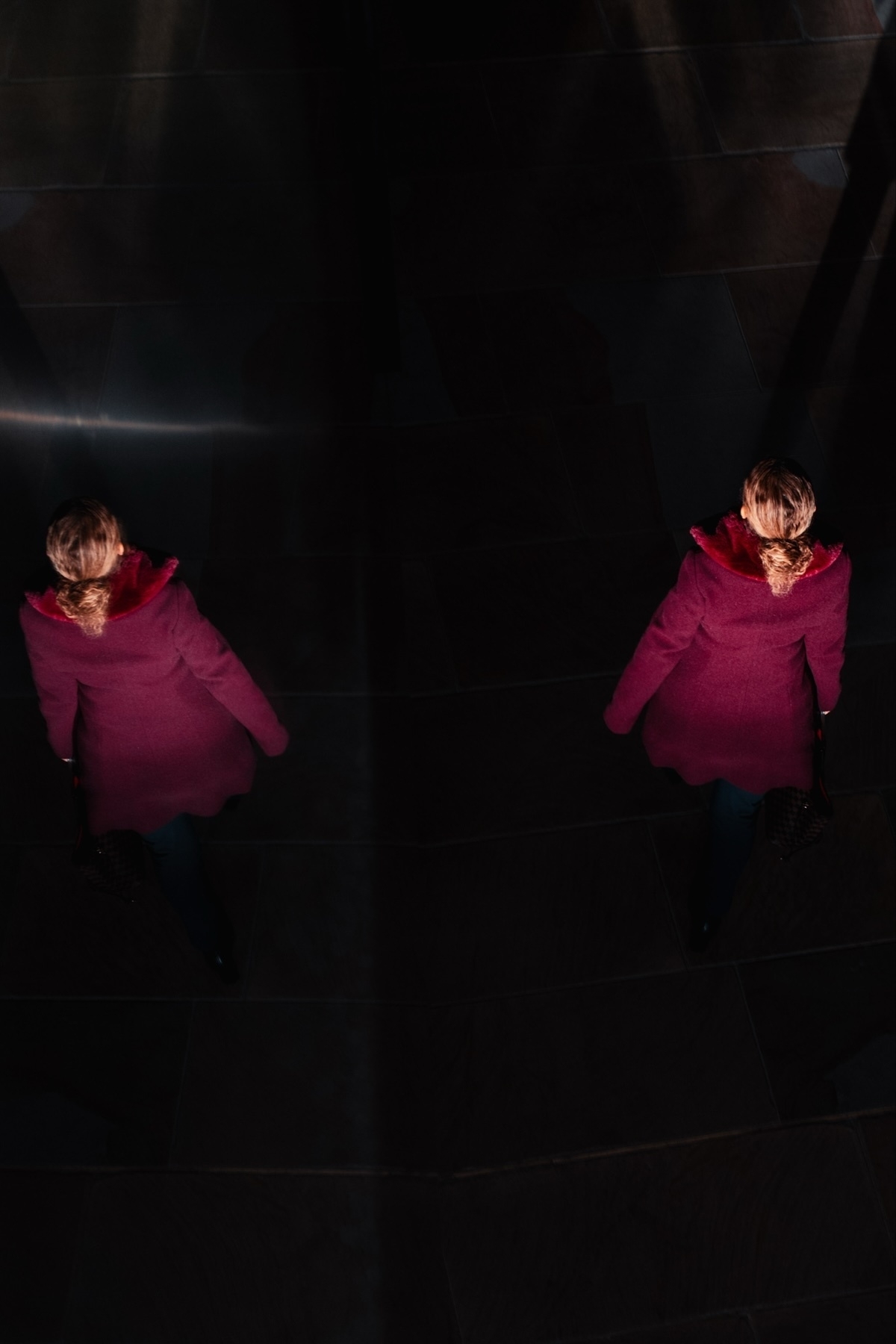 A person in a red coat is walking on a dark tiled floor, casting a shadow. The image is taken from above, with dramatic lighting highlighting the coat’s color and creating a reflection effect.