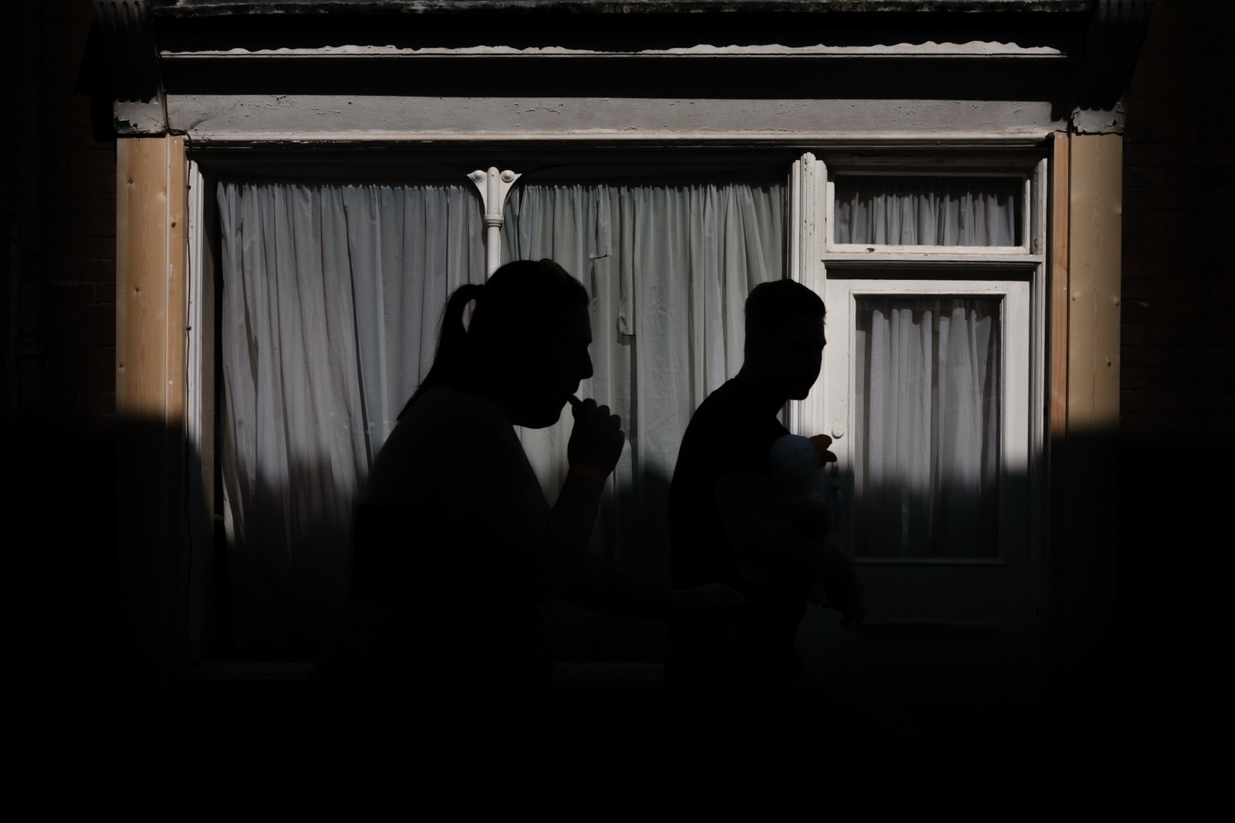 Silhouettes of two individuals walking in front of a window with closed curtains. The person in the foreground appears to be smoking, while the person behind is holding what seems to be a child or a large stuffed animal. The image is taken in lower