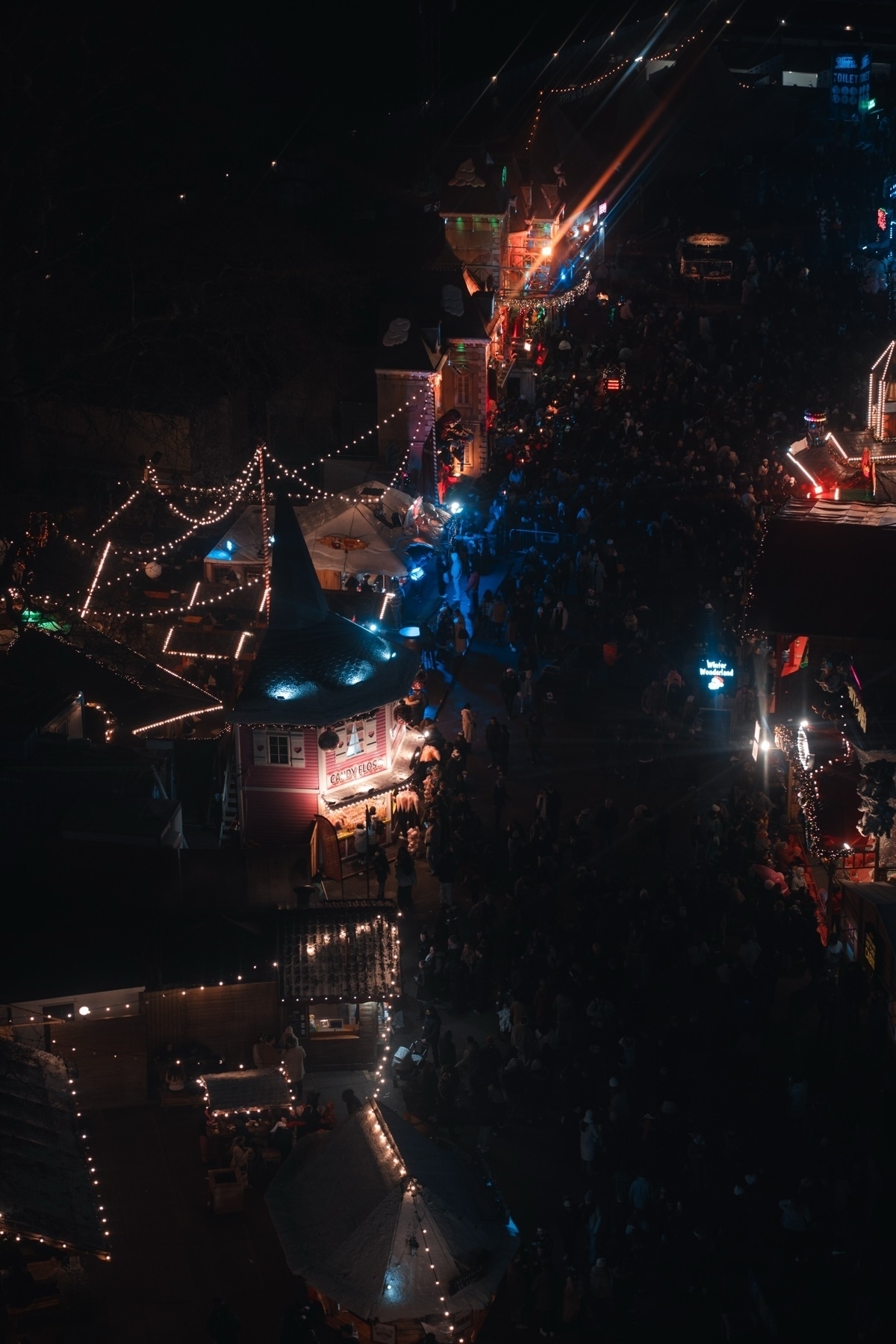 A vibrant nighttime carnival scene with festive lights and a large crowd. Stalls with candy and food are decorated with string lights, creating a lively atmosphere.