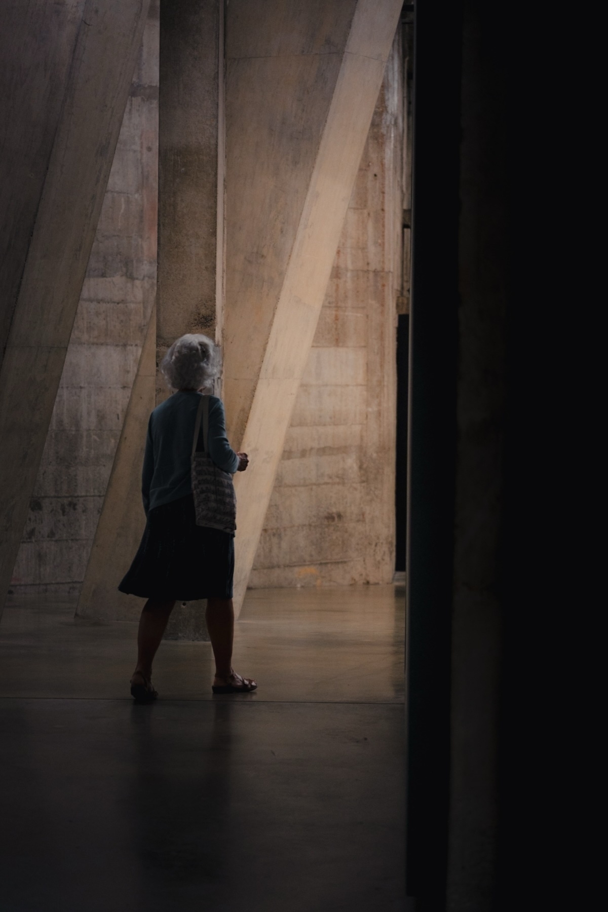 An elderly person with white hair, wearing a blue sweater and dark skirt, and holding a textured bag, walks through a dimly lit, industrial-looking space with large concrete pillars.