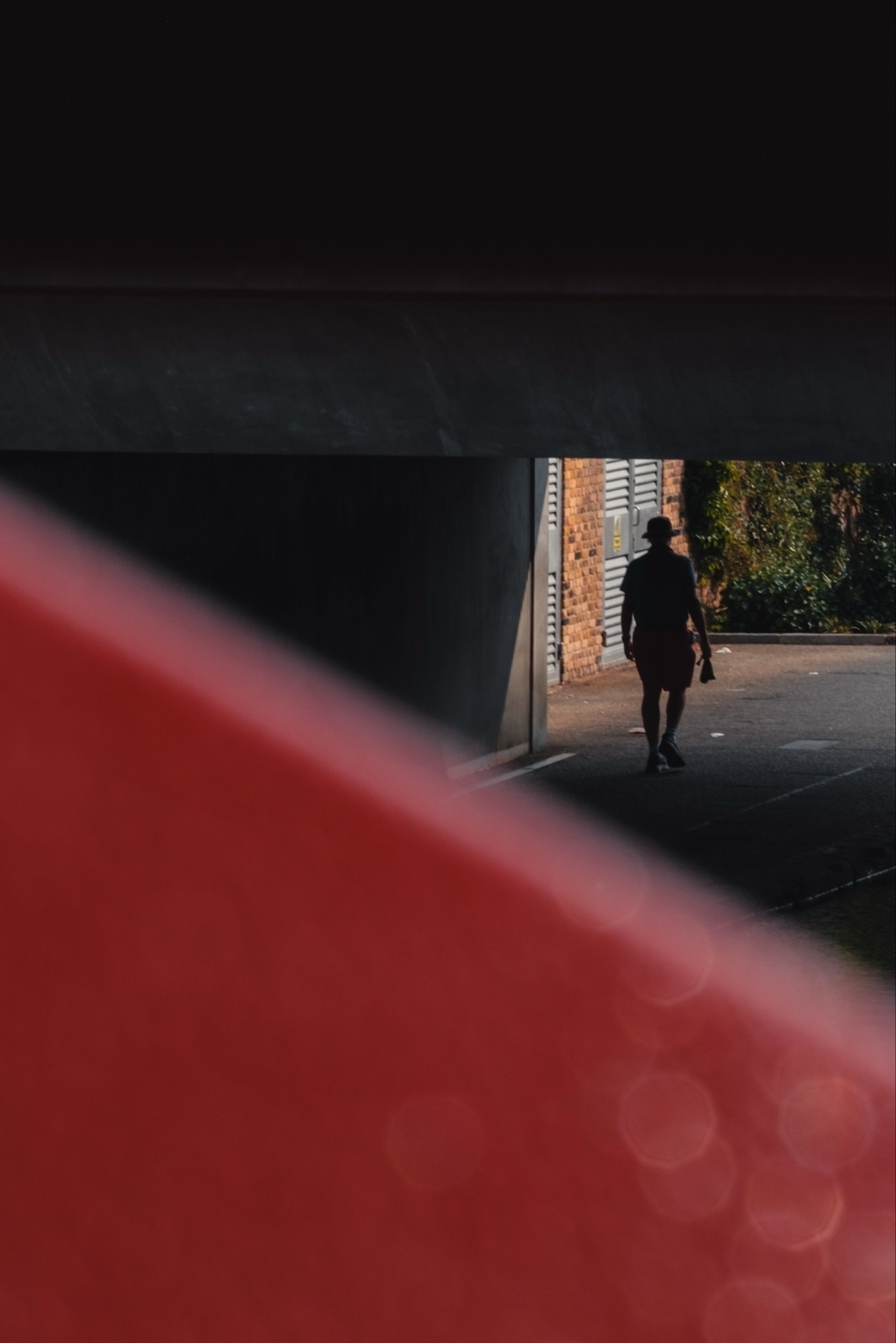 A person walking under a bridge, partially obscured by a red object in the foreground. The person is wearing a hat and shorts, and the background includes a brick wall and some foliage. The scene appears to be captured from a low angle with a