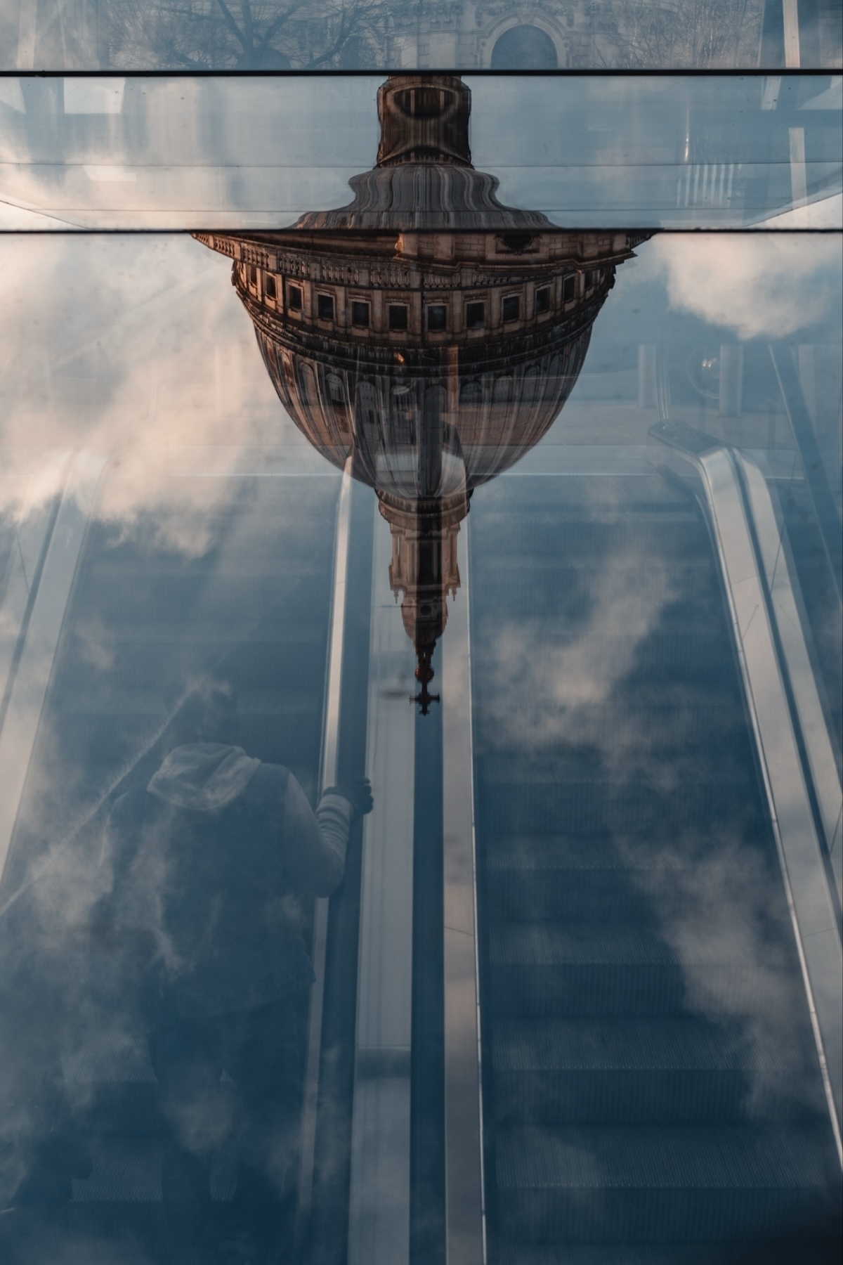 Reflection of a domed building on glass above an escalator. A person is ascending on the left side, with clouds visible in the reflection, creating a surreal composition.