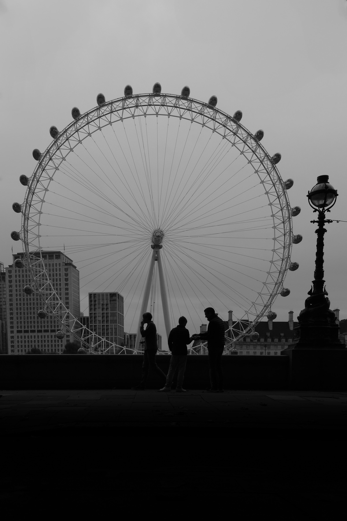 Silhouette of three people standing by a railing with the London Eye ferris wheel and city buildings in the background. The scene is set against a cloudy sky, creating a dramatic contrast.