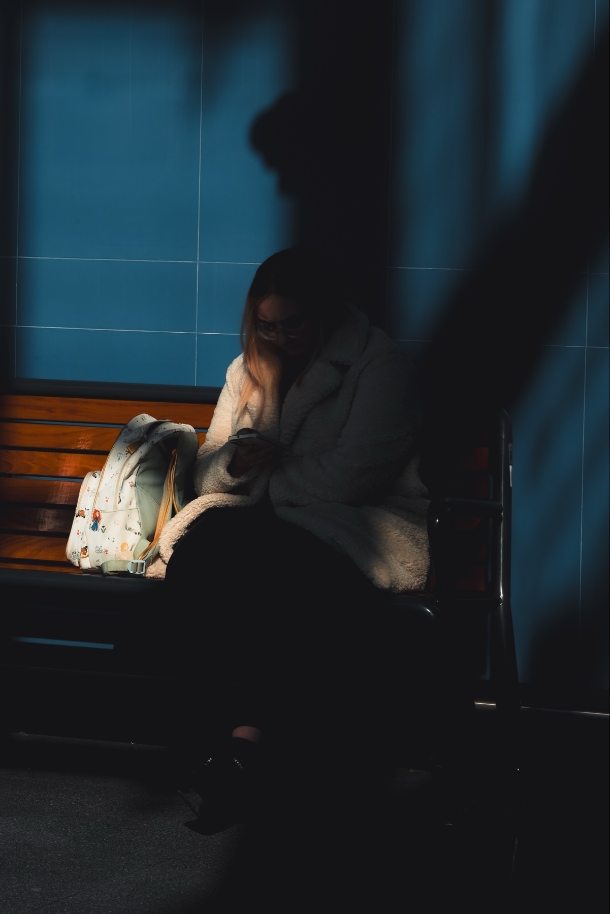 A person sits on a bench in a dimly lit area, wearing a fluffy coat and holding a phone. A colorful backpack rests beside them. Shadows create a dramatic effect against a blue-tiled wall.