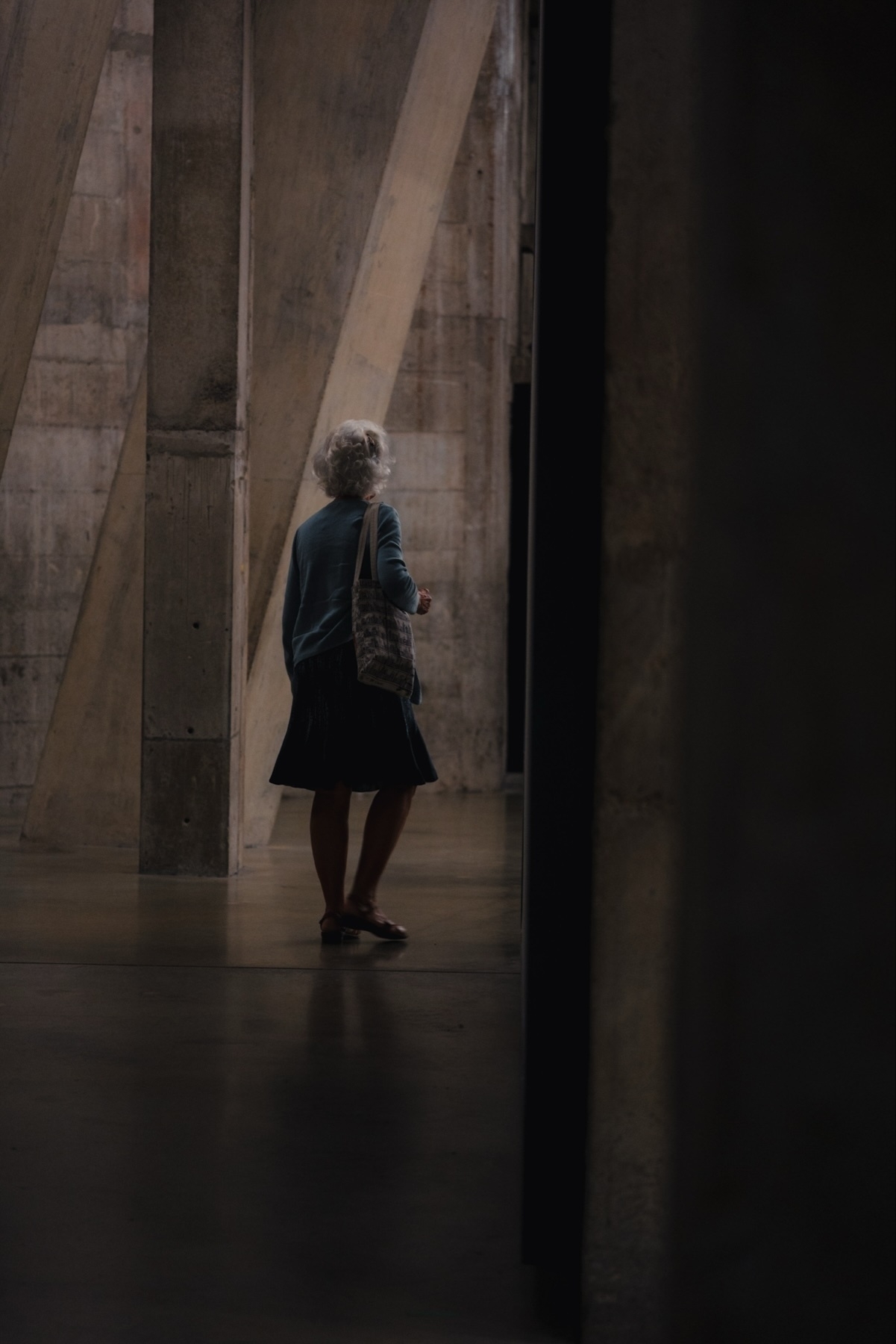 A person with gray hair wearing a blue sweater, dark skirt, and sandals, carrying a tote bag, walking through an industrial-style setting with large concrete support beams and dim lighting.