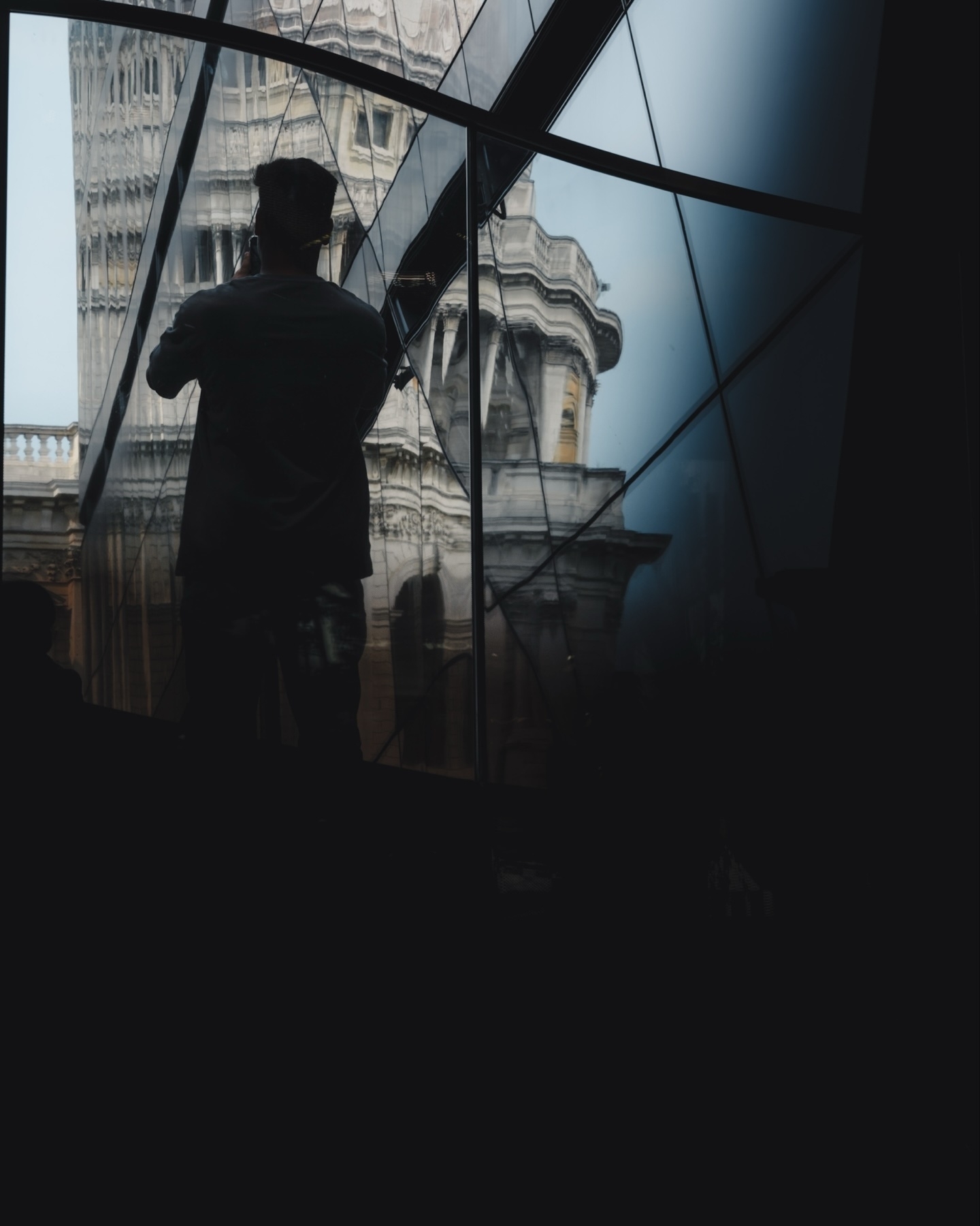 A person is silhouetted against a reflective, multi-panel glass surface, capturing an image of a historical building with distorted architecture visible in the reflection. The image has a dark, moody lighting aesthetic.