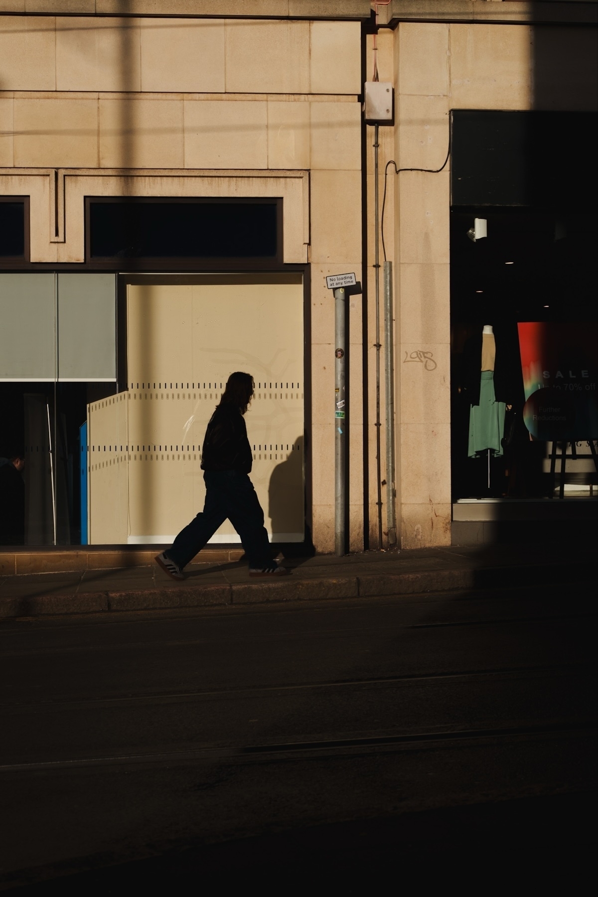 A silhouette of a person walking along a sidewalk is captured against a wall with a clear shadow. The building facade includes a window, a storefront sign, and a posted pole. The image conveys a contrast between sunlight and shadow, with a minimalist urban setting