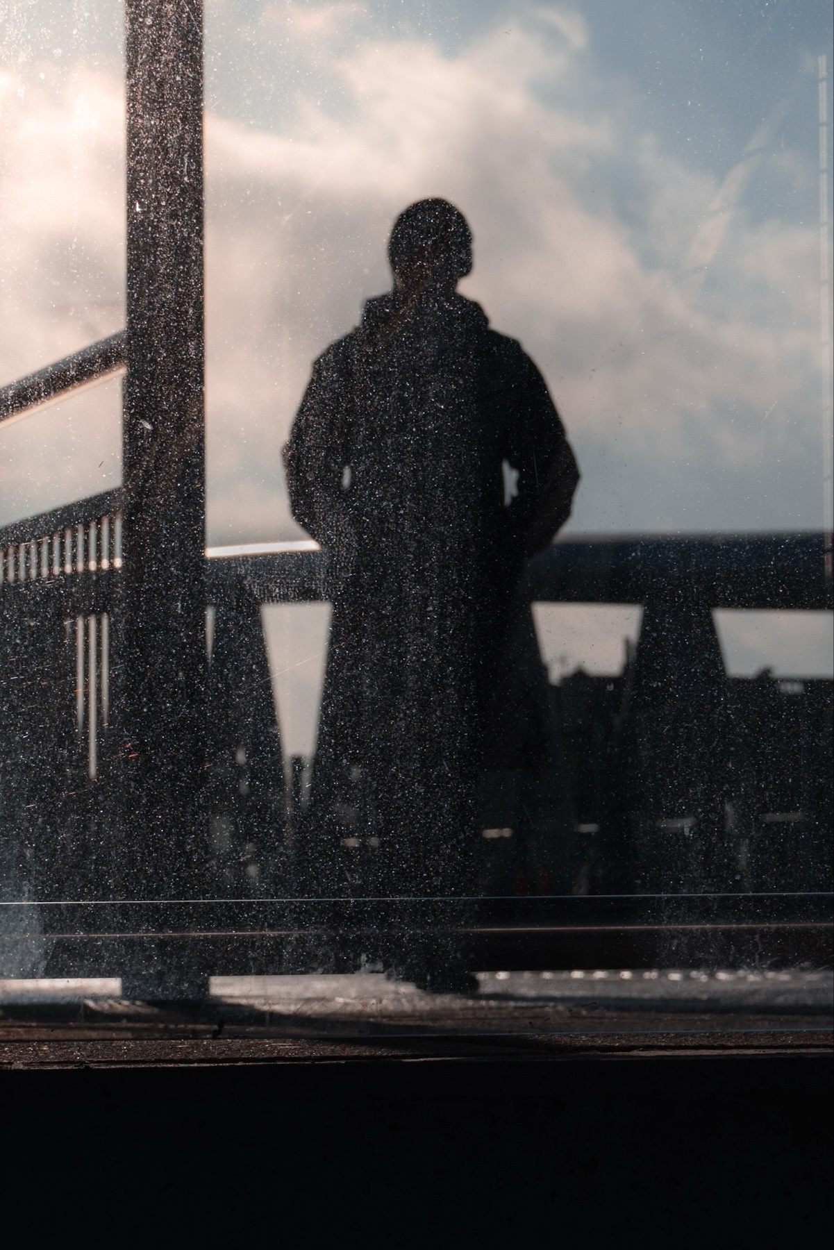 A silhouette of a person standing on a bridge is reflected in a dusty glass surface, with a cloudy sky in the background.