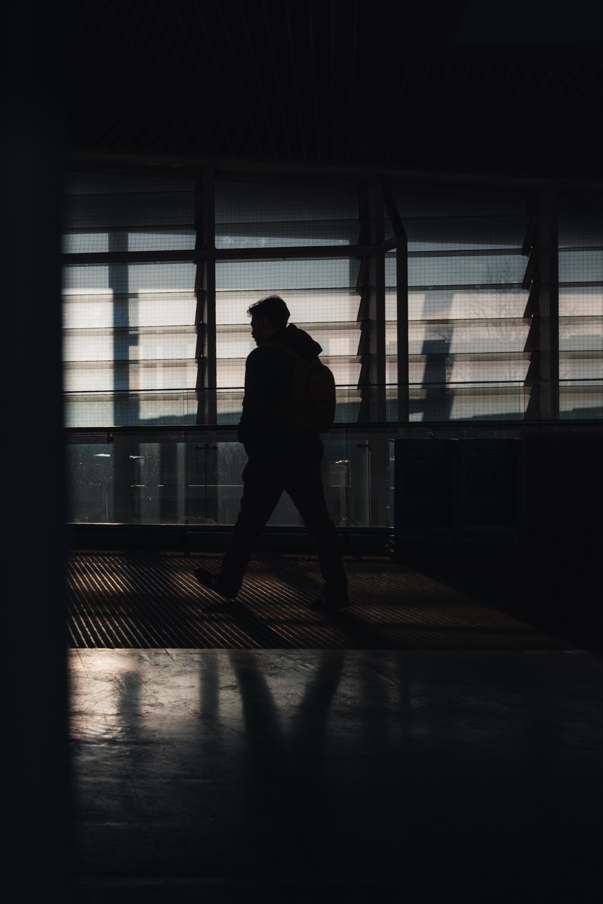 Silhouette of a person with a backpack walking indoors, set against large windows letting in soft, natural light.