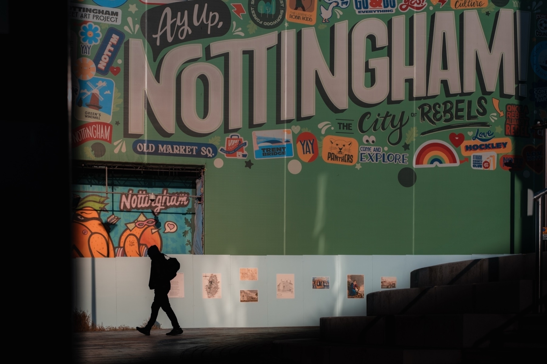 A silhouette of a person walking past a colorful mural in Nottingham. The mural features large text reading “Ay Up, Nottingham” and includes various local references, graphics, and phrases. The background is primarily green, with illustrations and vibrant details.