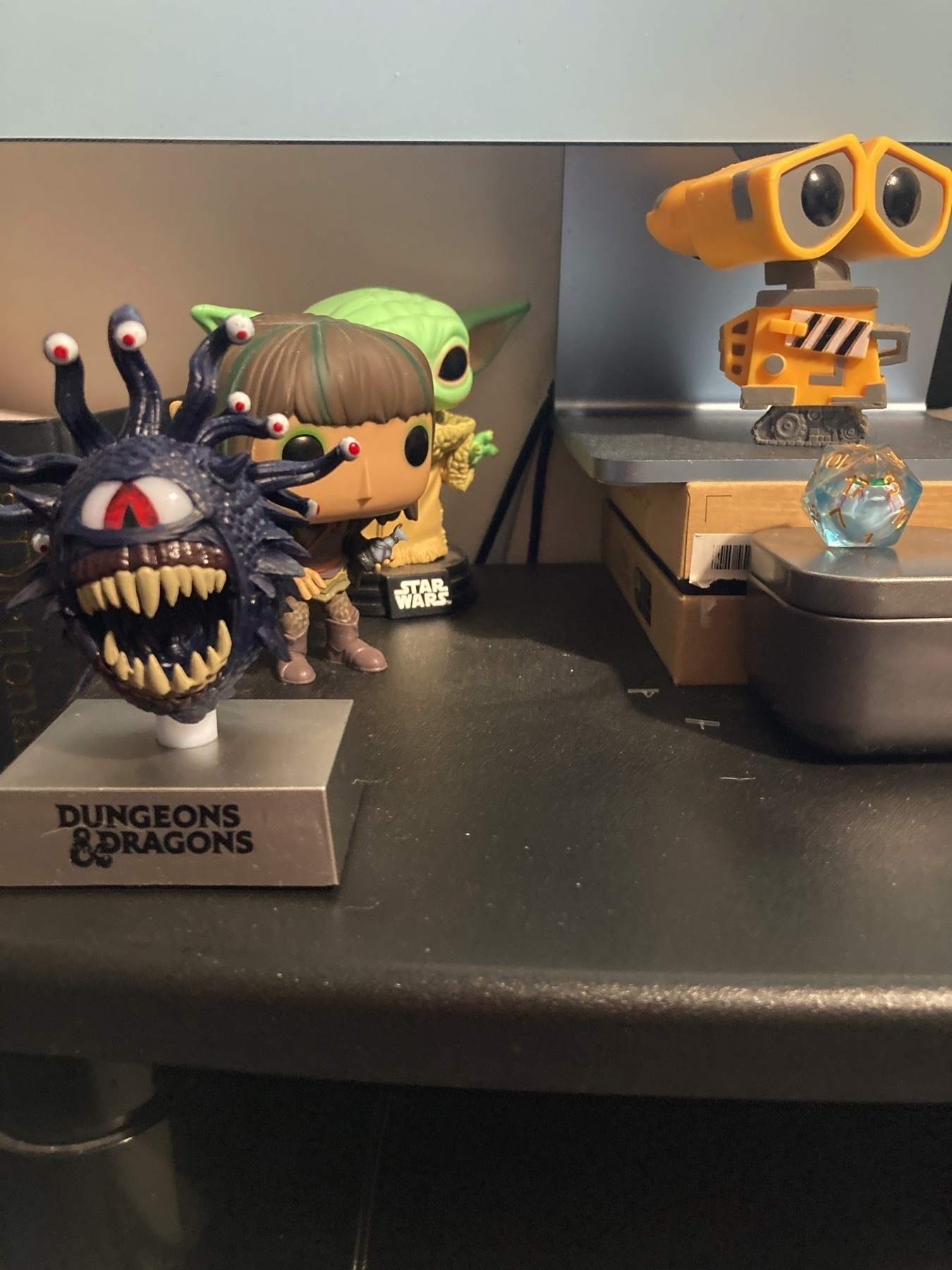 A selection of four figurines — a Beholder from Dungeons & Dragons, Rian from The Dark Crystal, Grogu from Star Wars, and Wall-E — and a die on top of a monitor stand, all positioned in front of a desktop computer.