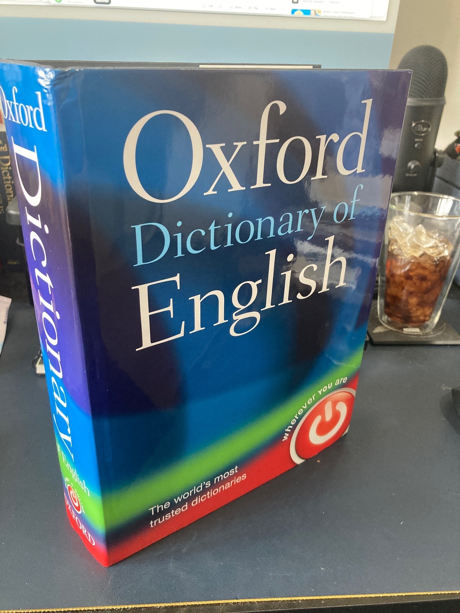 A large physical copy of the Oxford English Dictionary, standing on a desk in front of a keyboard and iMac.