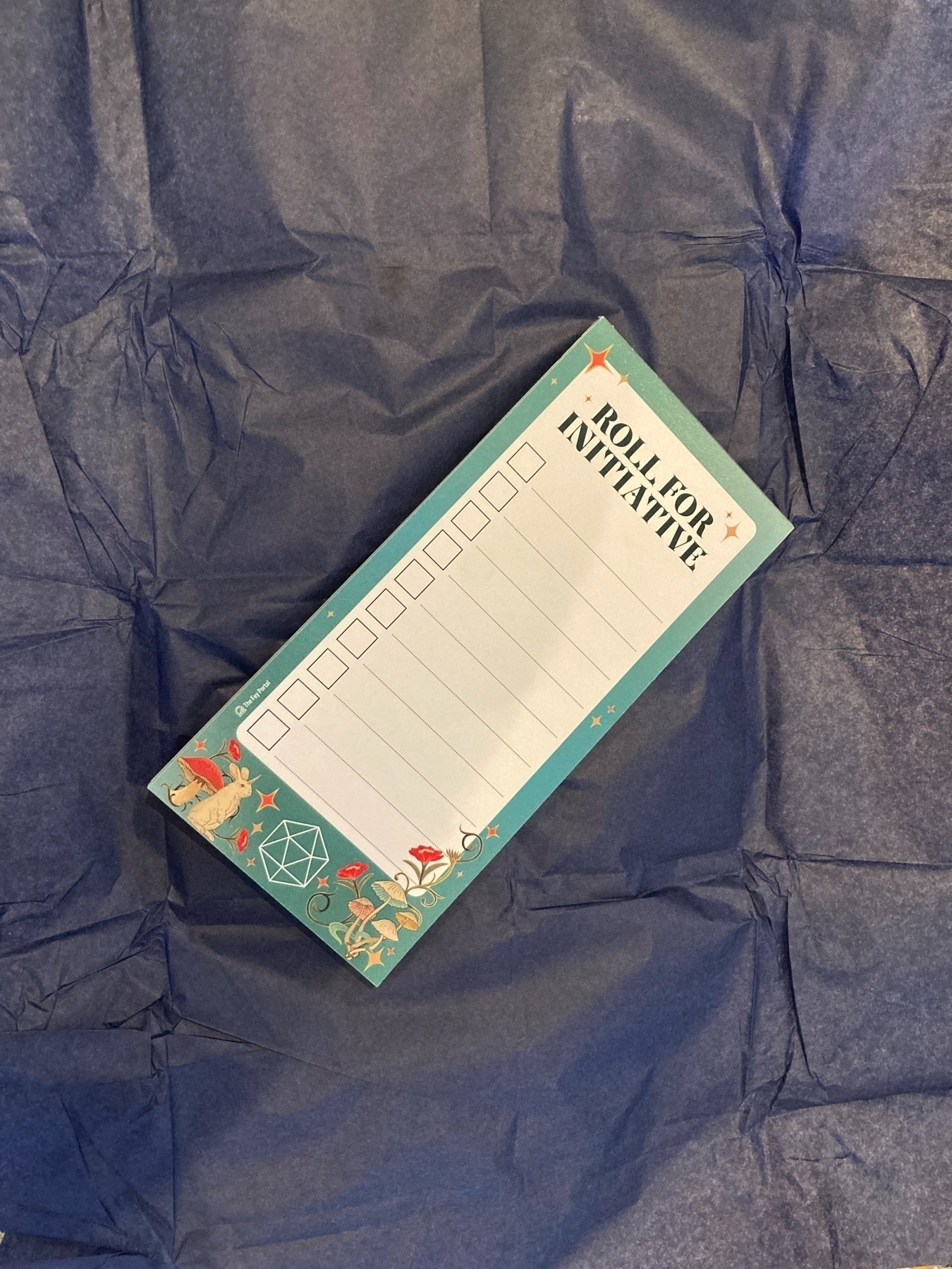 A notepad on top of blue tissue paper. The pad is formatted to easily list the names of characters in a role-playing game, specifically to track the order in which players may take their turn. The top of the page includes the words “roll for initiative” and there is a stylised border of green with a rabbit, flowers, mushrooms, and the outline of a 20-sided die.  