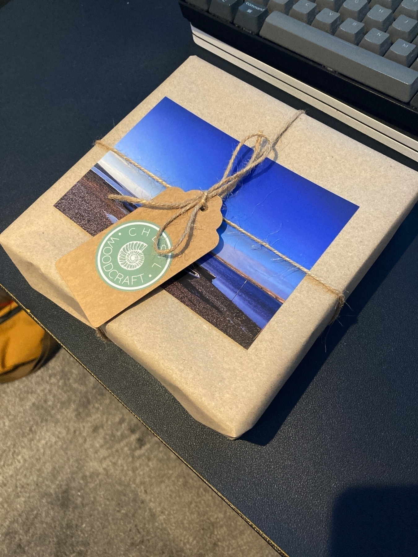 A wrapped parcel atop a table, in front of a keyboard. The wrapping is brown paper, with a postcard and tag attached via string. The sticker on the tag includes a seashell icon and the words “Chesil Woodcraft”.
