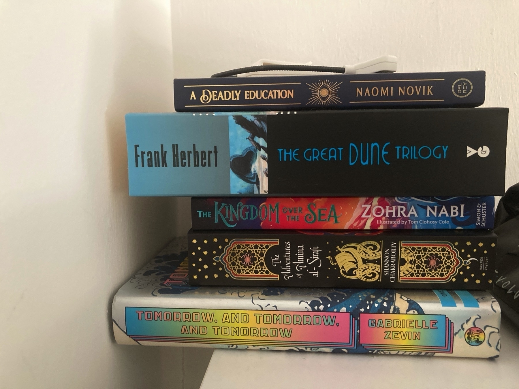 A stack of four books, in the corner of a hallway, with a small book-reading torch atop them. The books are: A Deadly Education, by Naomi Novik; The Great Dune Trilogy, by Frank Herbert; The Kingdom Over the Sea, by Zohra Nabi; The Adventures of Amina al-Sirafi, by Shannon Chakraborty; and Tomorrow, and Tomorrow, and Tomorrow, by Gabrielle Zevin.