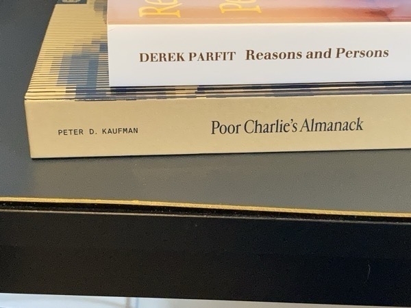 A close capture of two books atop a mat, on a table. The top one is “Reasons and Persons” by Derek Parfit, whilst the bottom one is “Poor Charlie’s Almanack” by Peter D. Kaufman.