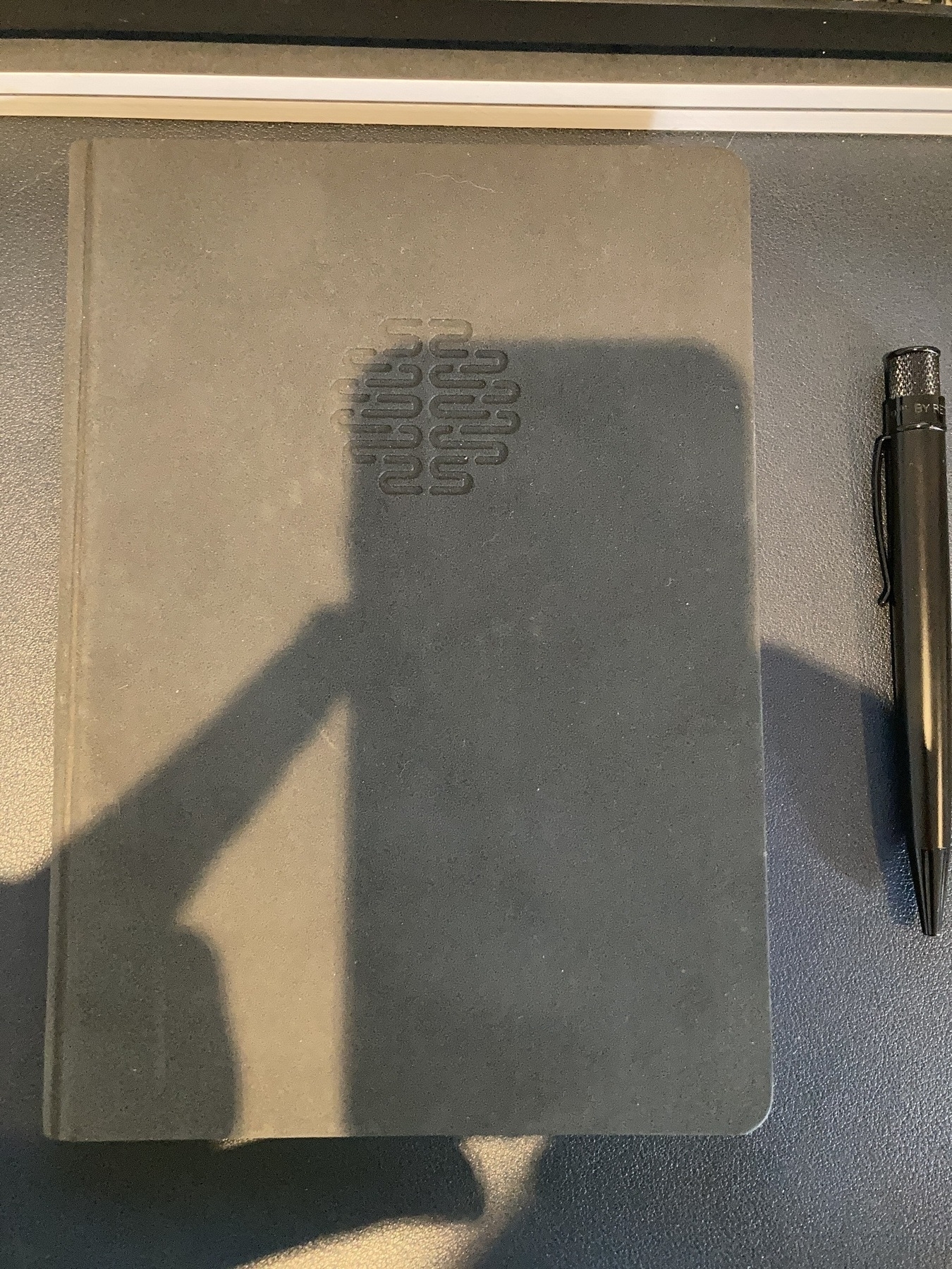 A dark notebook from above, with the shadow of my iPhone covering part of it. It is the Theme Journal notebook, with the brainwave symbol embossed in the upper-centre position of the front cover.