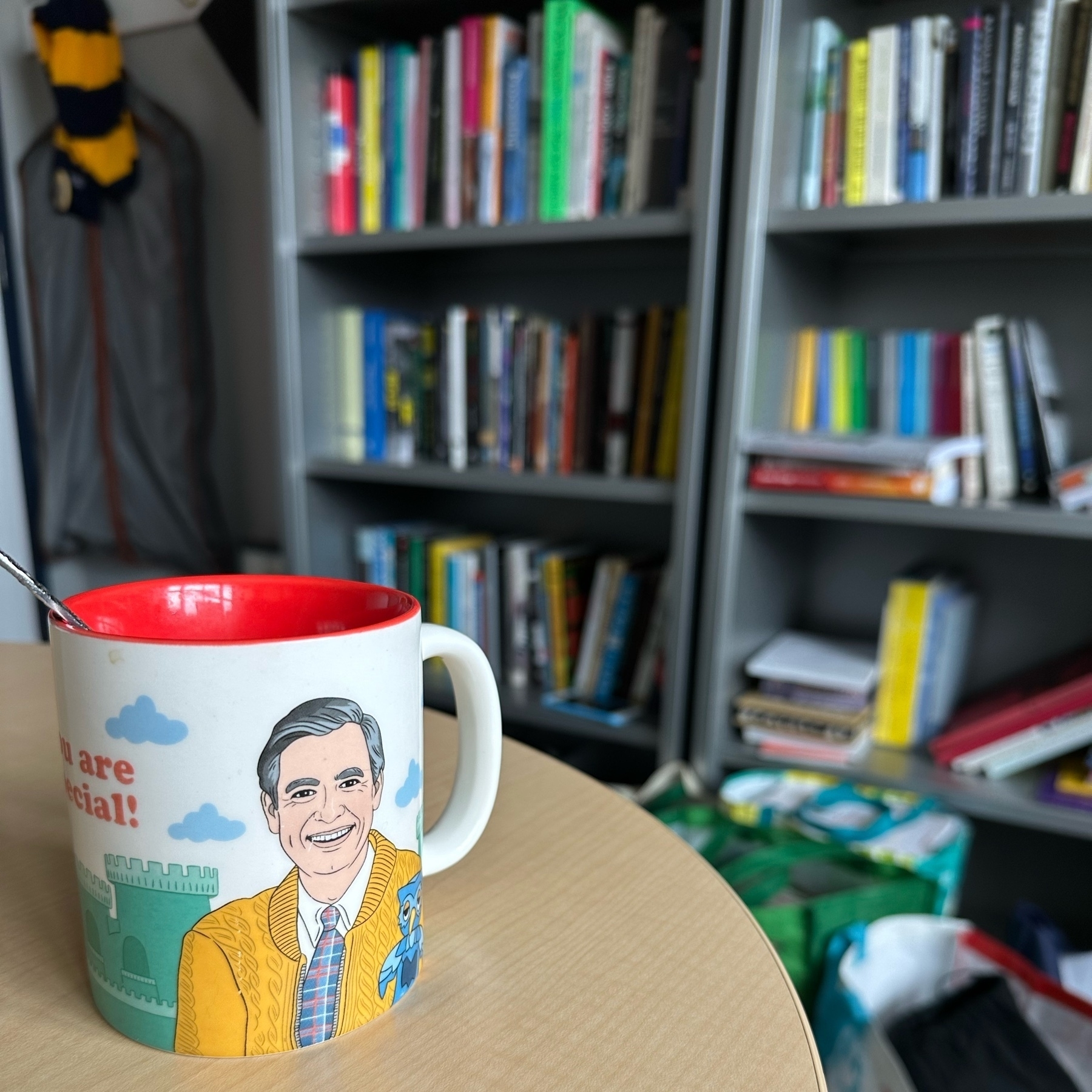 Coffee mug featuring a rendering of Fred Rogers (aka Mr. Rogers) on a desk with bookshelves in the background. 