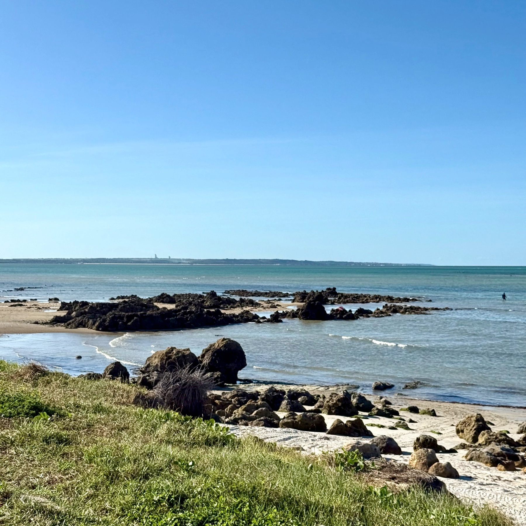 Uma paisagem costeira tranquila com pedras na areia e o mar ao fundo sob um céu azul.