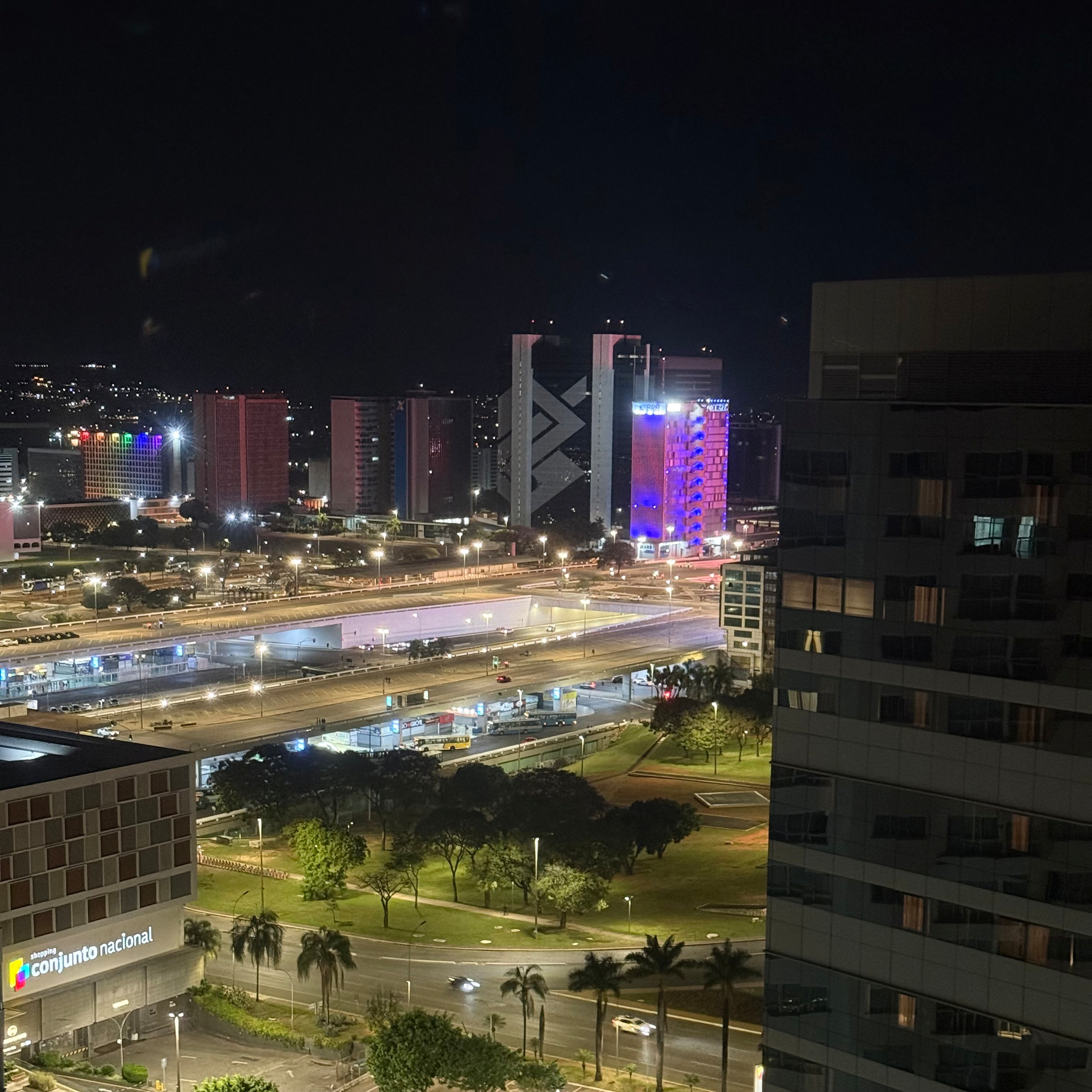 Vista noturna de uma área urbana com edifícios iluminados, uma via expressa movimentada e áreas verdes com árvores e palmeiras.