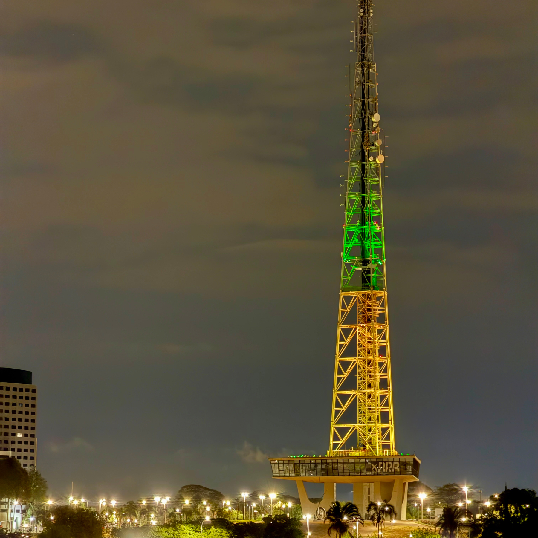 Uma torre de transmissão iluminada com as cores verde e amarela, destaque em uma paisagem urbana durante a noite.