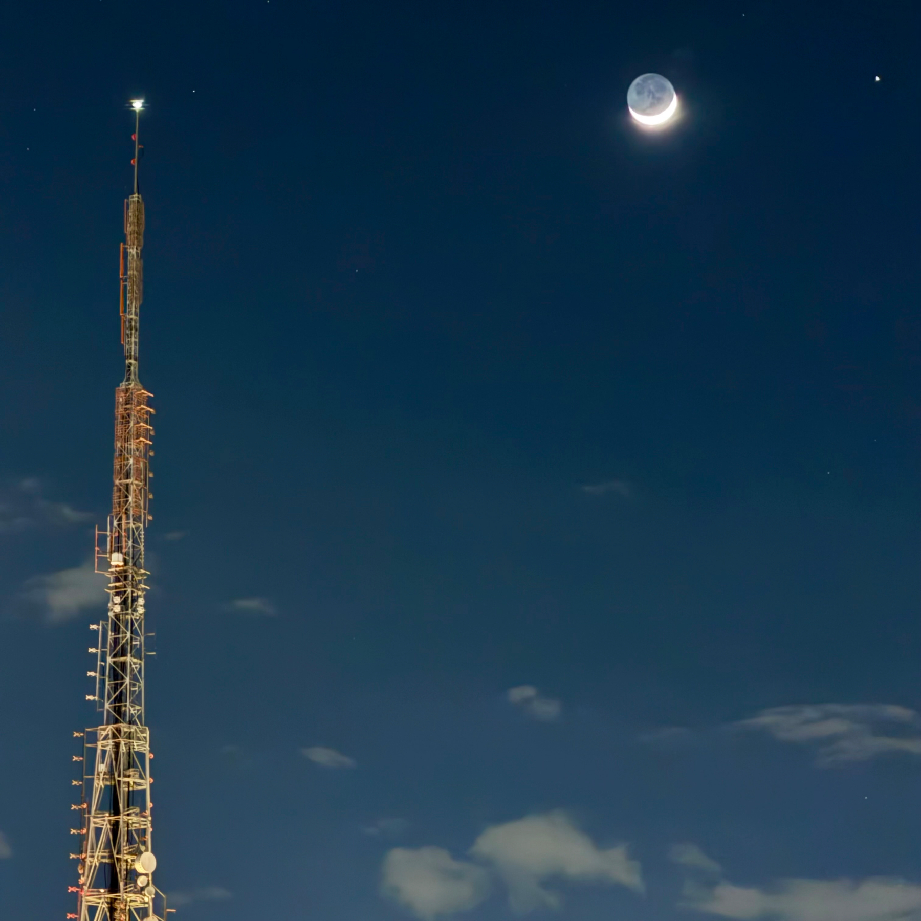 Uma torre de transmissão está iluminada à noite sob um céu escuro com a lua crescente e algumas estrelas visíveis.