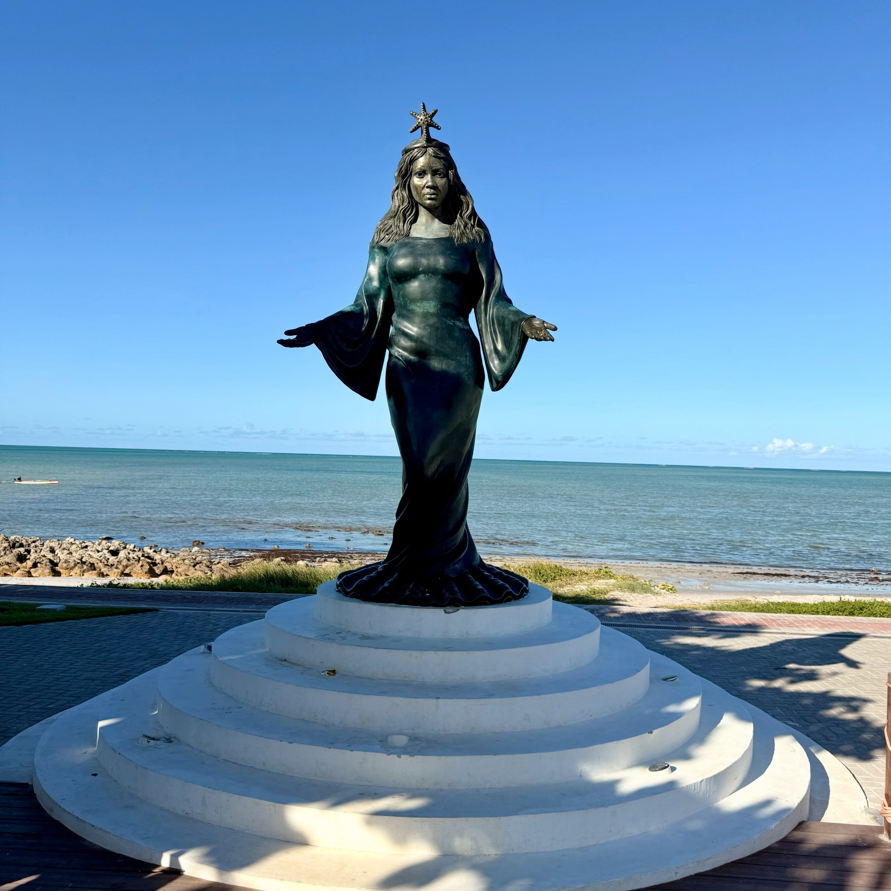 Uma estátua de uma figura feminina de braços abertos está sobre um pedestal em frente ao mar.