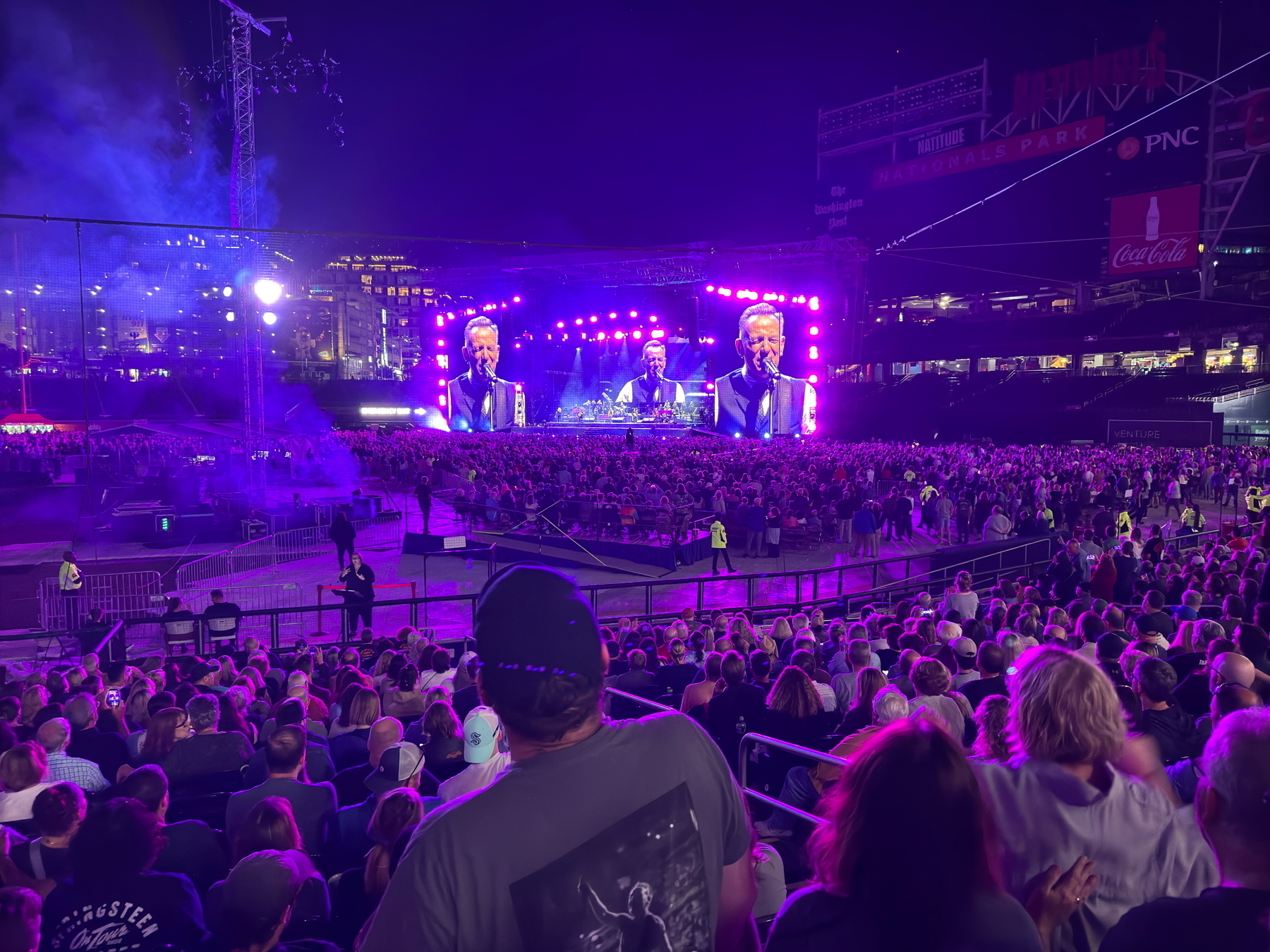 Auto-generated description: A large crowd is attending a Bruce Springsteen show with a brightly lit stage and screens displaying Springsteen.