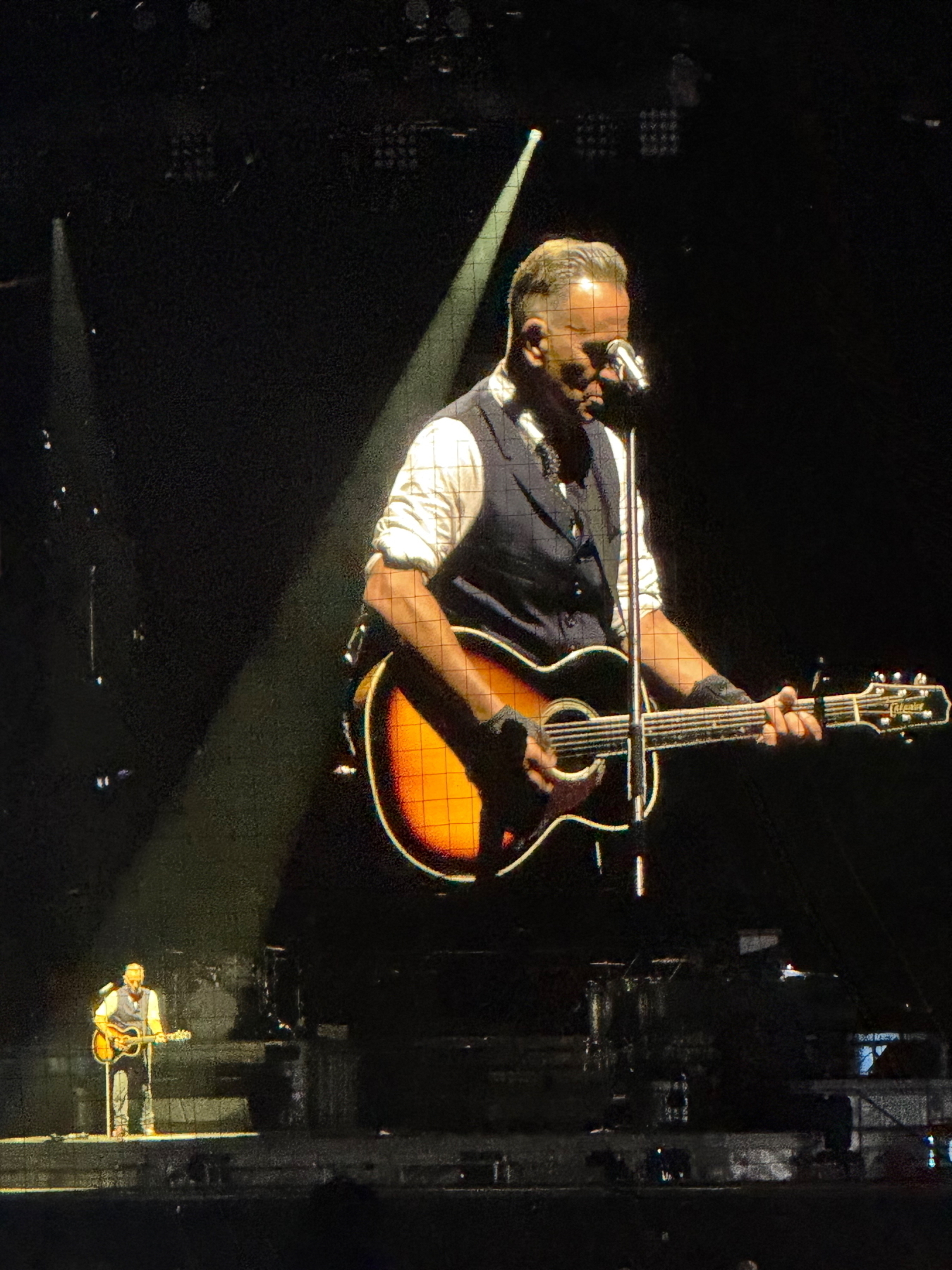 Auto-generated description: Bruce Springsteen playing an acoustic guitar and singing on stage, with a large screen projecting his image behind him.