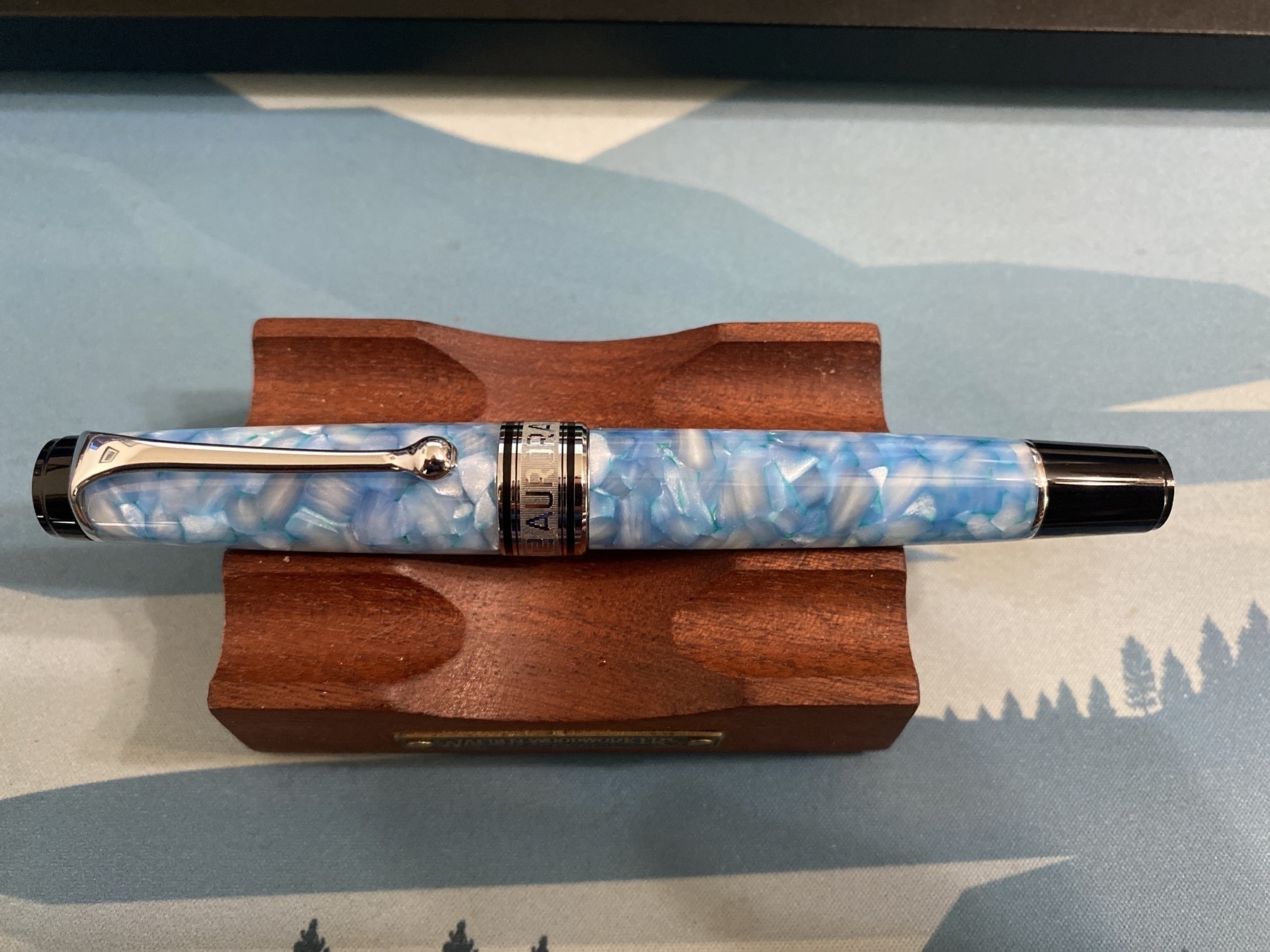 An Aurora Optima 365 Celeste fountain pen, capped and resting on a wooden pen rest.