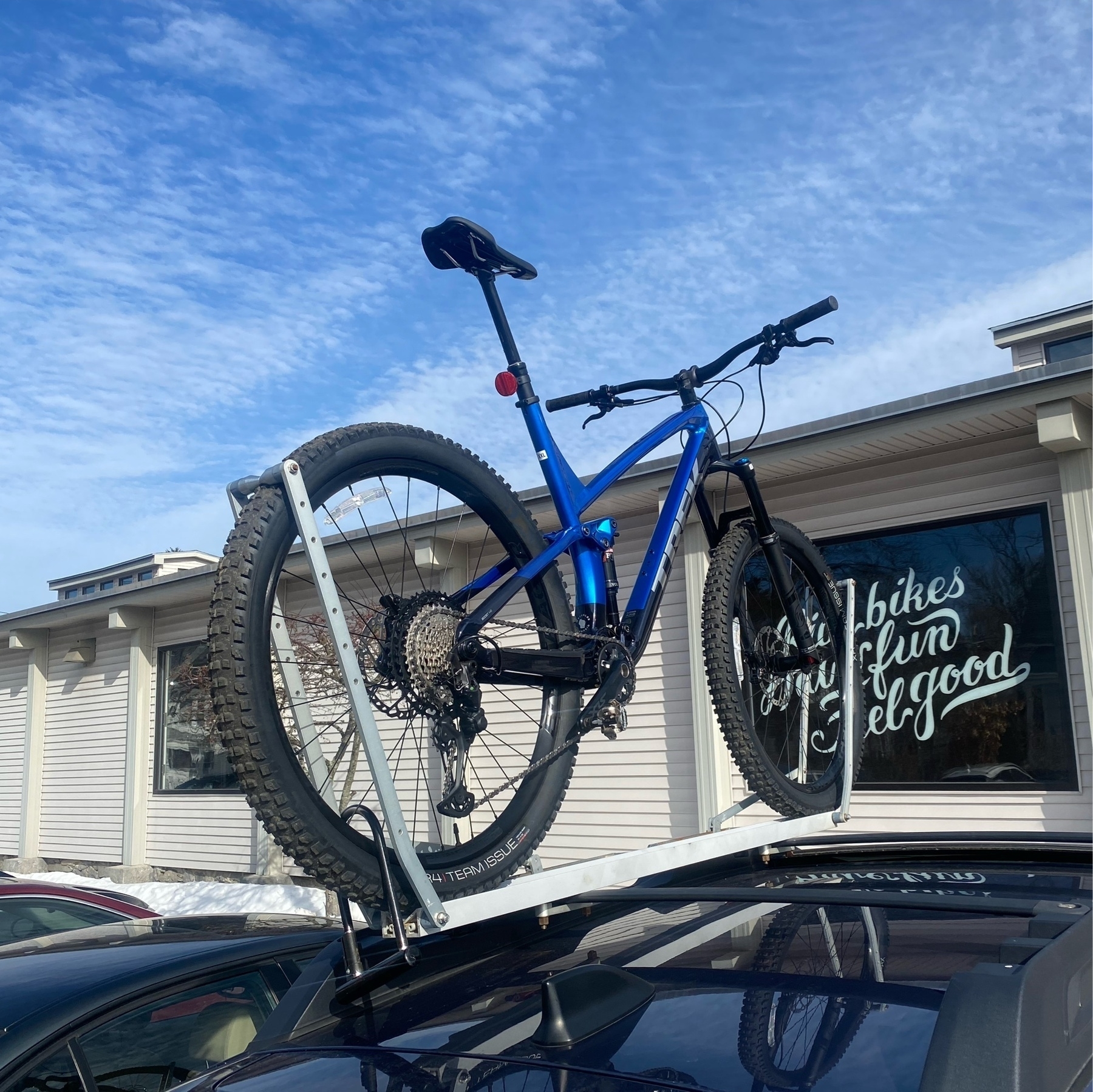 a shiny blue Trek Fuel mountain bike on a roof rack with a bright blue sky and the top of a building in the background.
