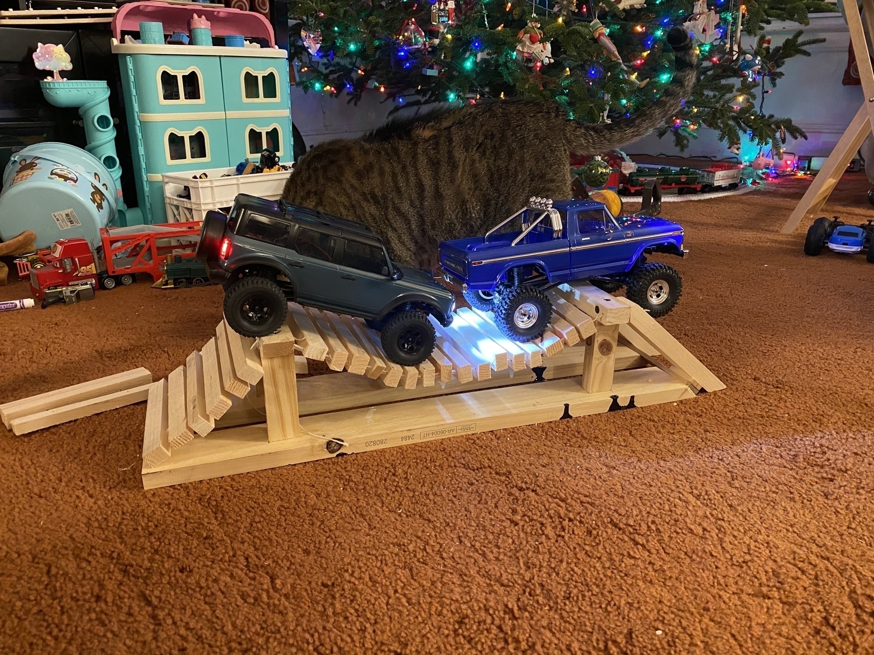 Two 1/18th scale rc crawler trucks on a homemade mini rope bridge. In the background is a car, kid’s toys, a Christmas tree, and a Christmas train.