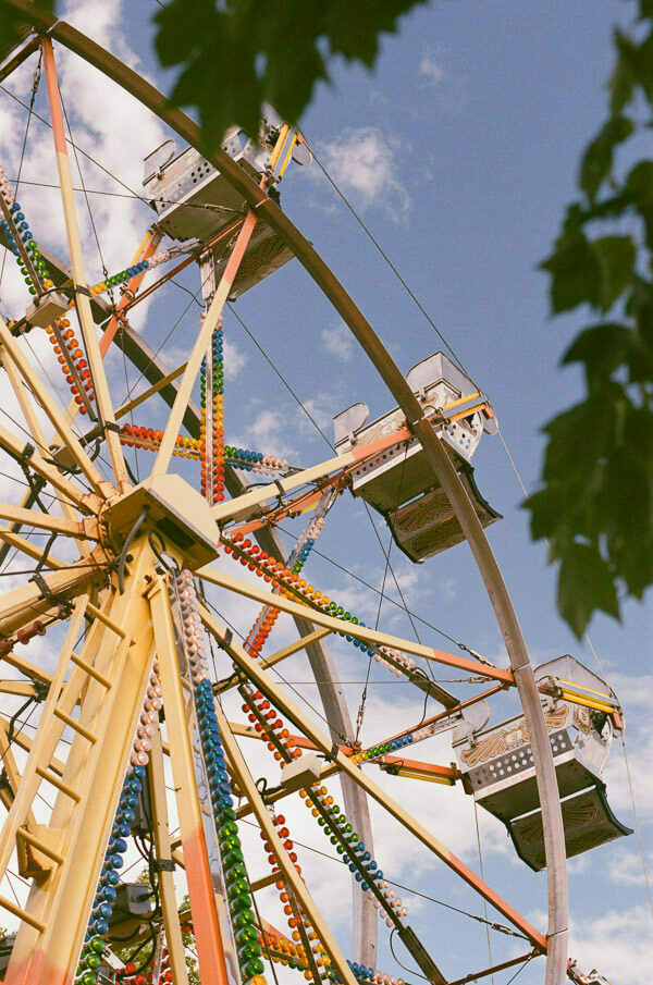 A close-up shot of a Ferris wheel at a fair, showing colorful cabins decorated with strings of lights. The sky is clear with a few clouds, and the top of a tree can be seen at the bottom of the frame.