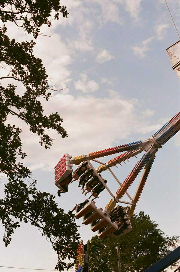 An action shot of a fairground ride in motion, with chairs suspended on chains swinging out. The ride is multicolored, and trees frame the top of the image against a backdrop of a blue sky with soft clouds.