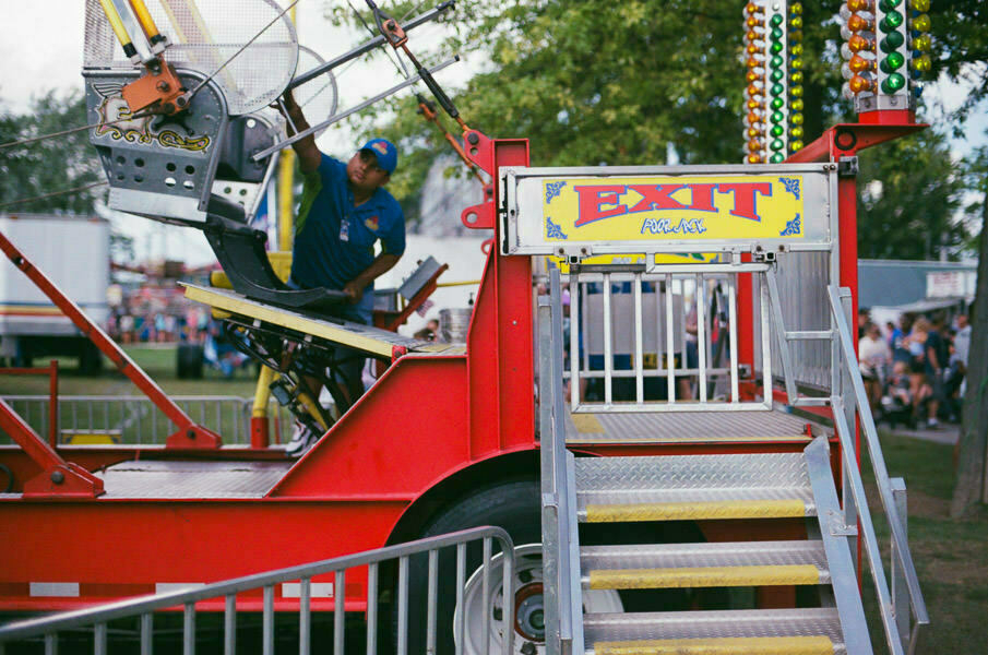 A carnival ride operator standing at the exit gate of a ride, which is painted red and yellow. The ride is not in motion, and there's a crowd of fairgoers in the background behind a metal barrier.