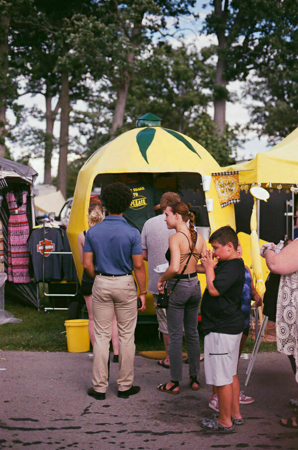 A queue of people waiting at a lemon-shaped food trailer. The booth is bright yellow with a green tip, resembling a lemon, and is set against a busy fairground background.