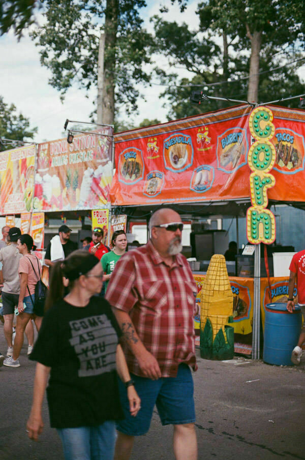 A bustling fairground scene with a food stall in the foreground advertising 'Corn Dogs,' 'Nachos,' 'Tacos,' and more. People are walking by in various directions, and there's a colorful banner overhead.