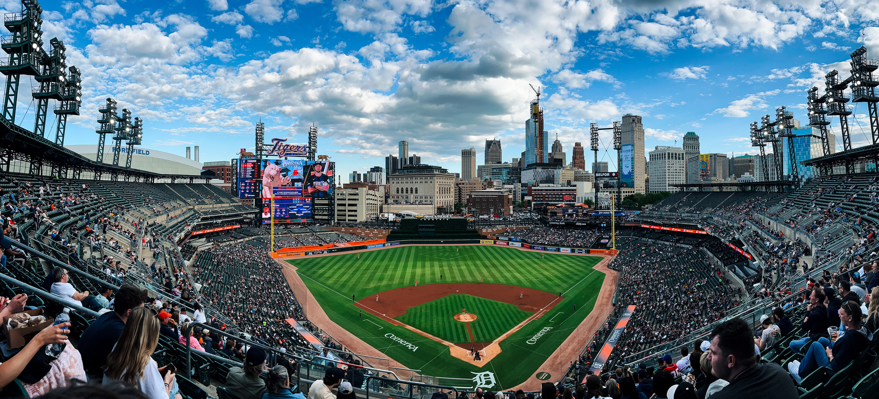 A panorama photograph of Comerica Park in Detroit, Michigan, on a partially cloudy day as the Tigers play the Houston Astros. Photograph is taken from behind home plate showing the Detroit skyline in the distance. 