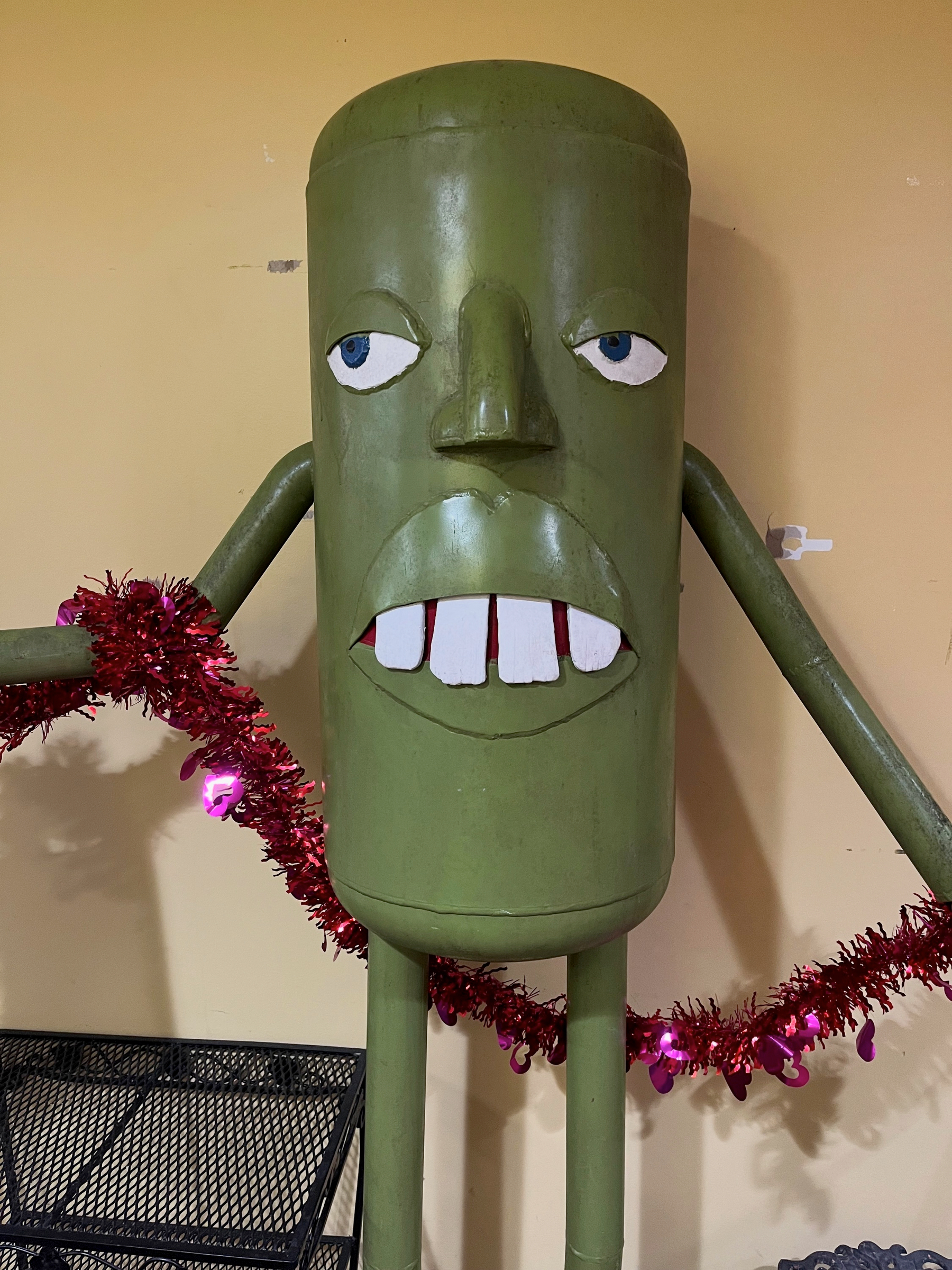 A green cylindrical figure with a cartoonish face, long arms, and legs is holding a red tinsel garland against a beige wall.